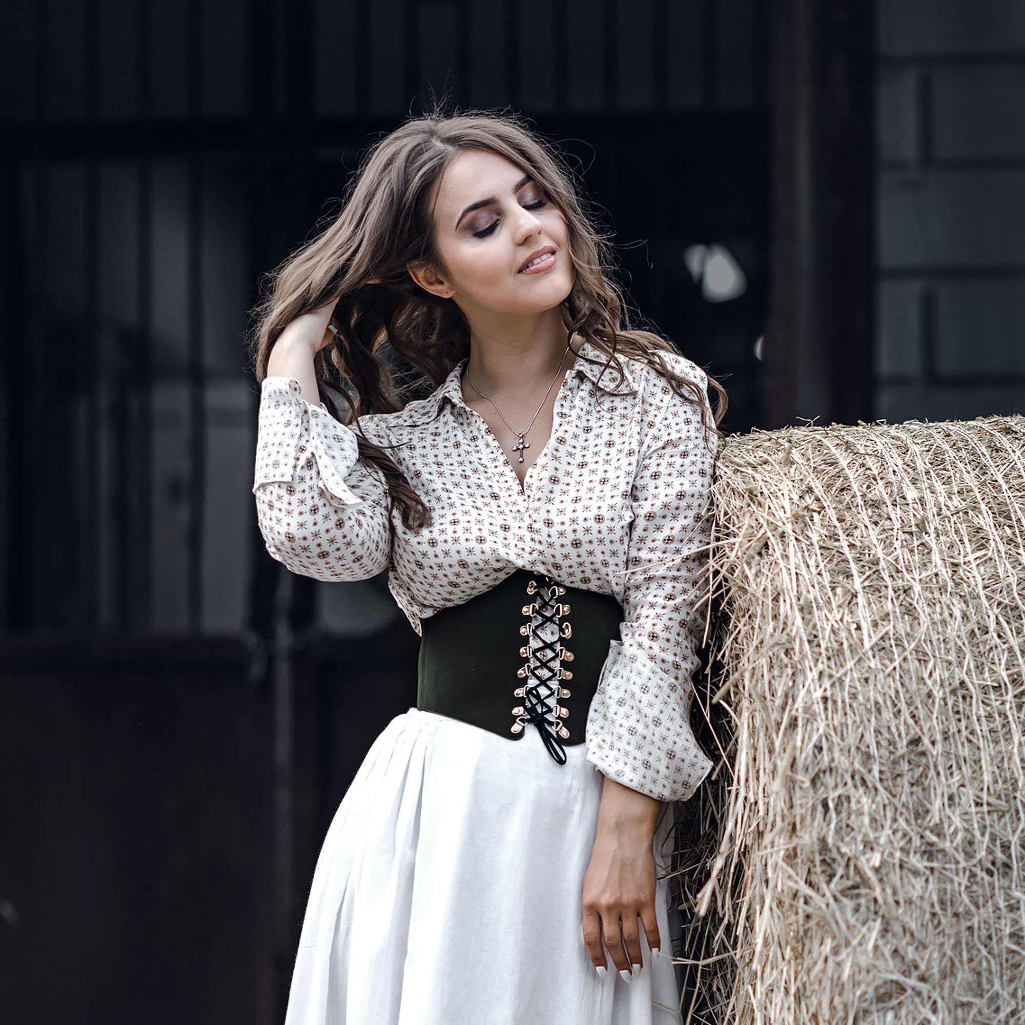 Портрет девушки с длинными каштановыми волосами летом на конном дворе возле стога сена на фоне конюшни