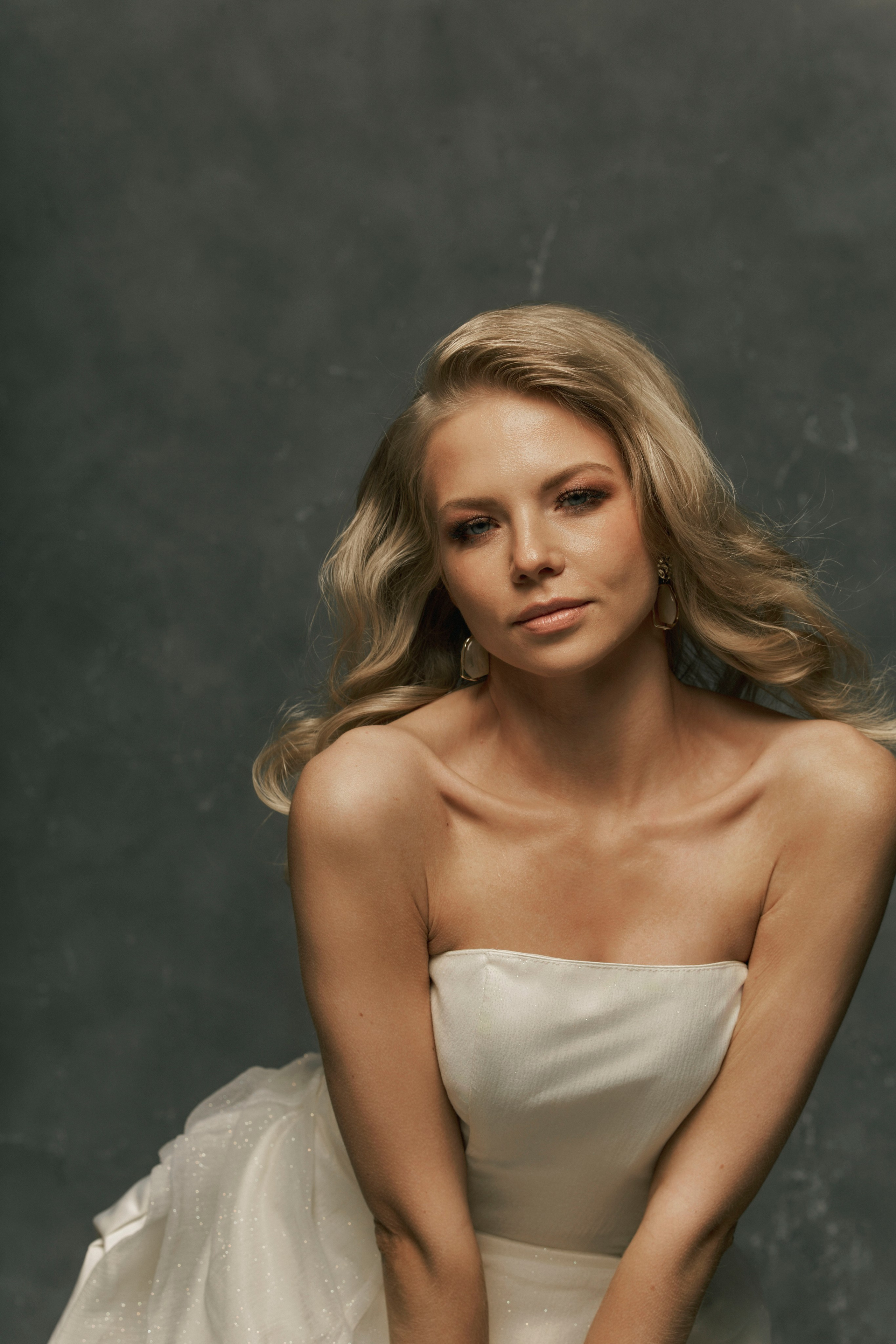 Две женщины в вечерних платьях на фоне гардеробной стойки с одеждой. Одна из них стоит в ярко-красном платье с объемным декором в виде роз, а другая сидит в воздушном белом платье. Интерьер оформлен в темных тонах с элементами драпировки. На переднем плане заметны книги и туфли на высоких каблуках.