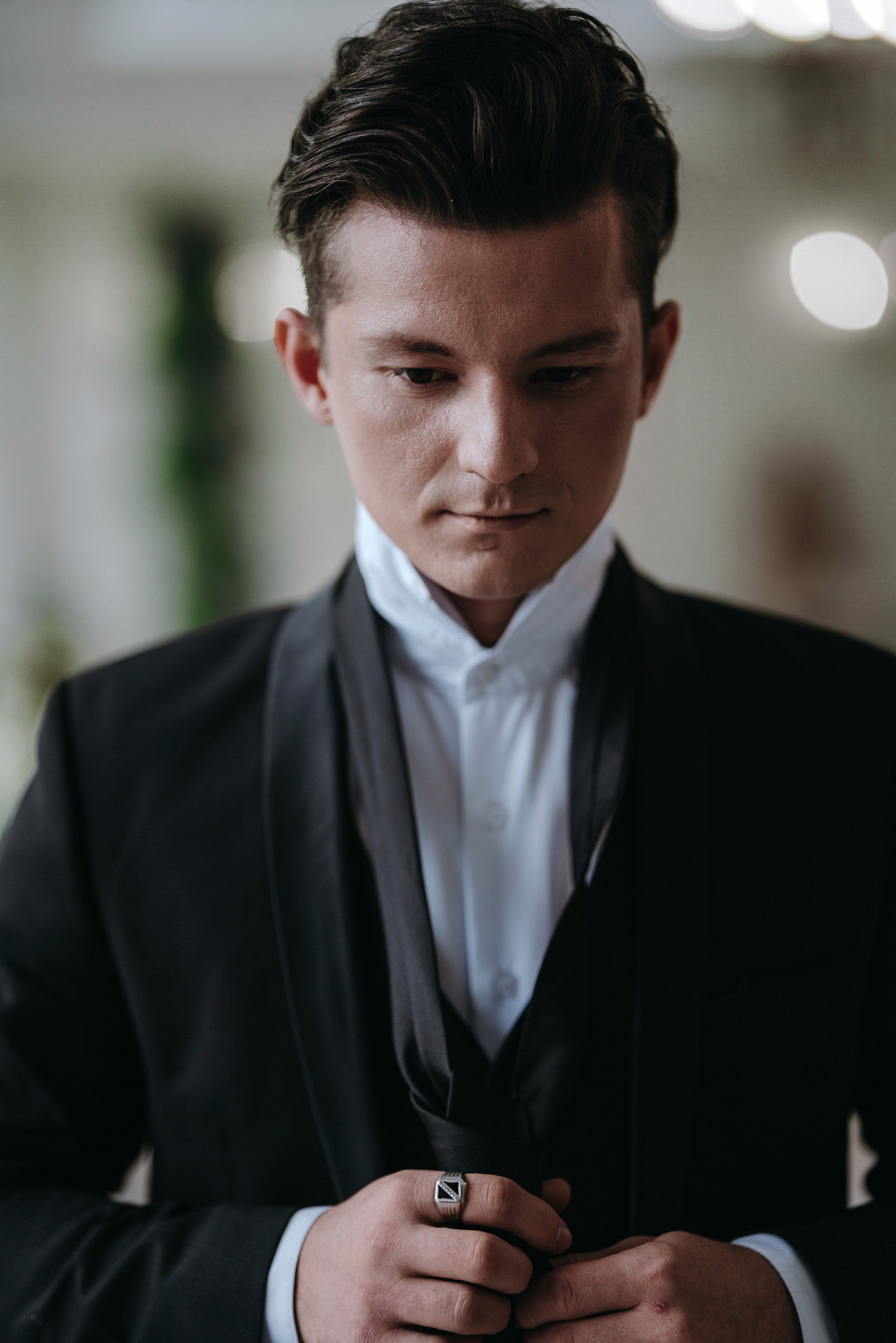 Молодой мужчина в чёрном смокинге и белой рубашке сосредоточенно поправляет детали костюма, стоя на фоне с тёплым рассеянным светом