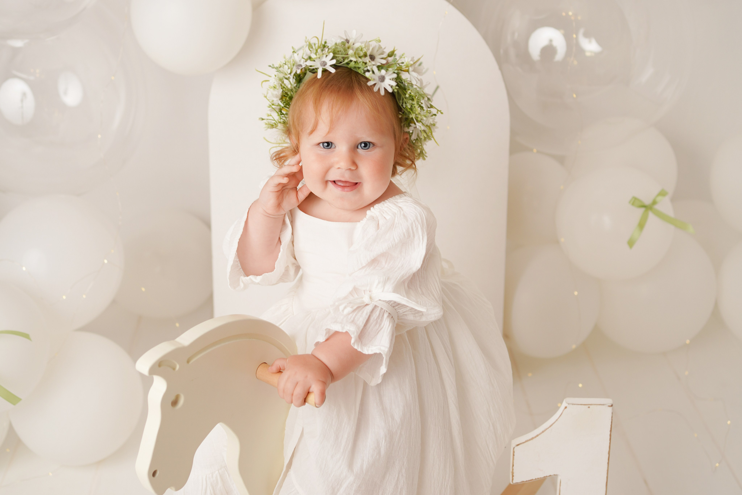 Малышка в белом муслиновом платье и цветочном веночке катается на игрушечной лошадке.