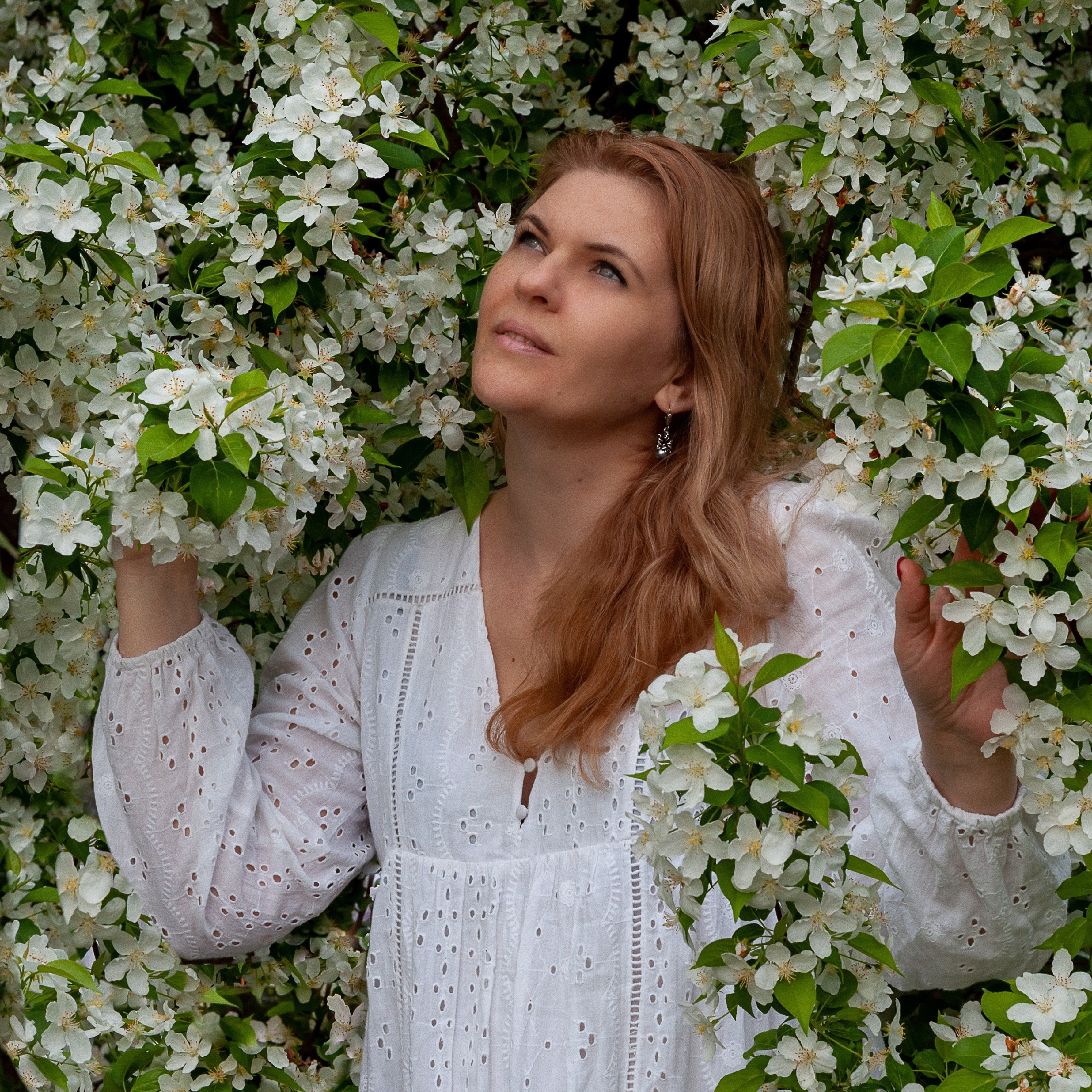 Женщина в белом платье позирует среди цветущих яблоневых деревьев в весеннем саду, наслаждаясь ароматом природы.