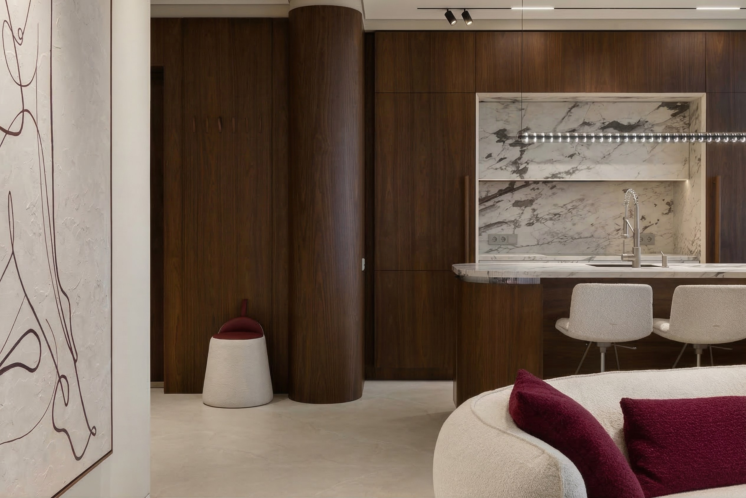 Фрагмент интерьера в ЖК ONYX Deluxe: радиусная стена и шкафы из темного дерева, кухонный остров с мраморным фартуком и светлый пуф. Дизайн Валерии Котович.