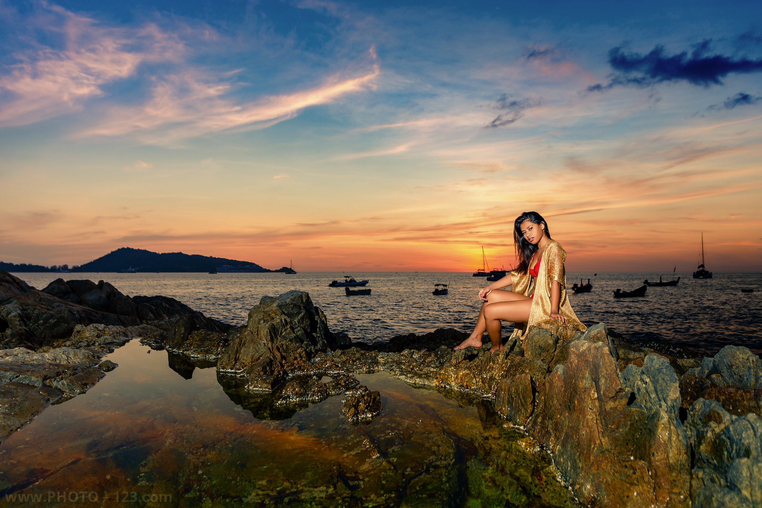 Sunset beach portrait photography, woman sitting on coastal rocks at golden hour, tropical seascape with boats, dramatic sky, lifestyle fashion photoshoot by the ocean, professional travel photographer portfolio