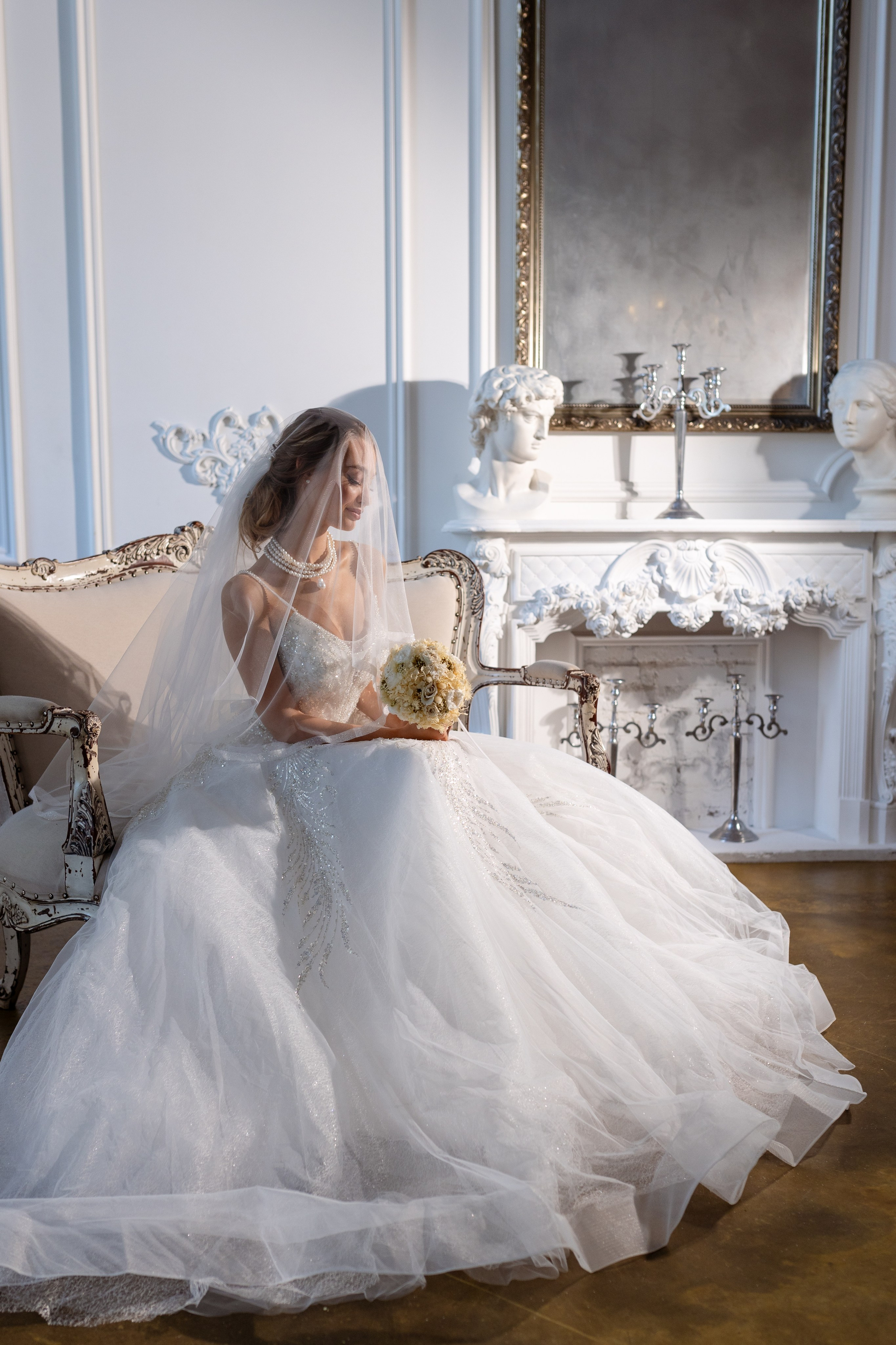 Невеста сидит на диване на фоне белоснежного столика.