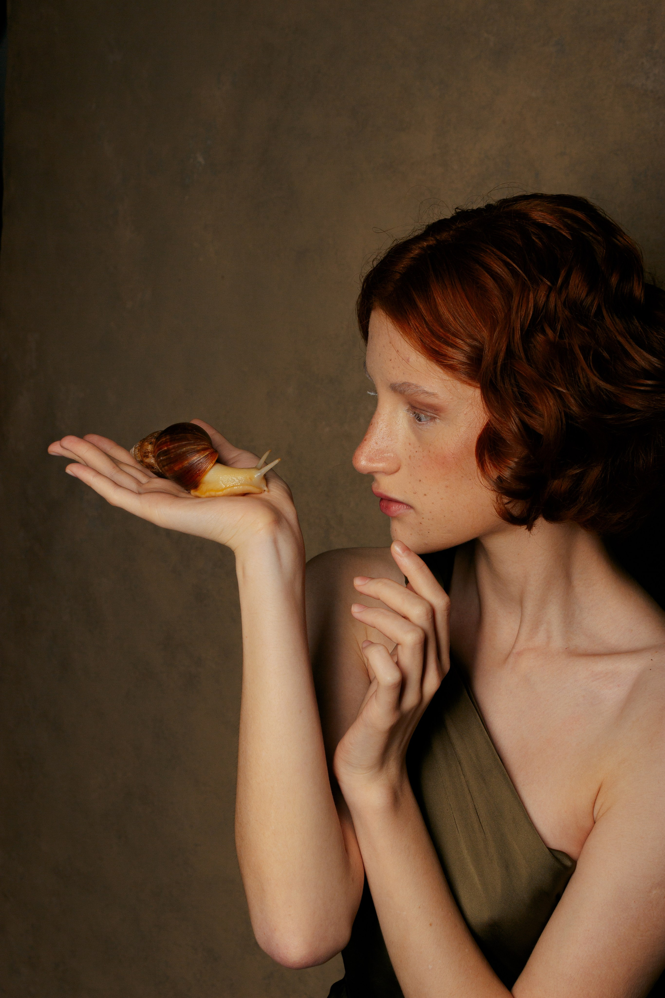 молодая рыжая девушка держит на ладошке большую улитку Ахатину и удивленно на нее смотрит 