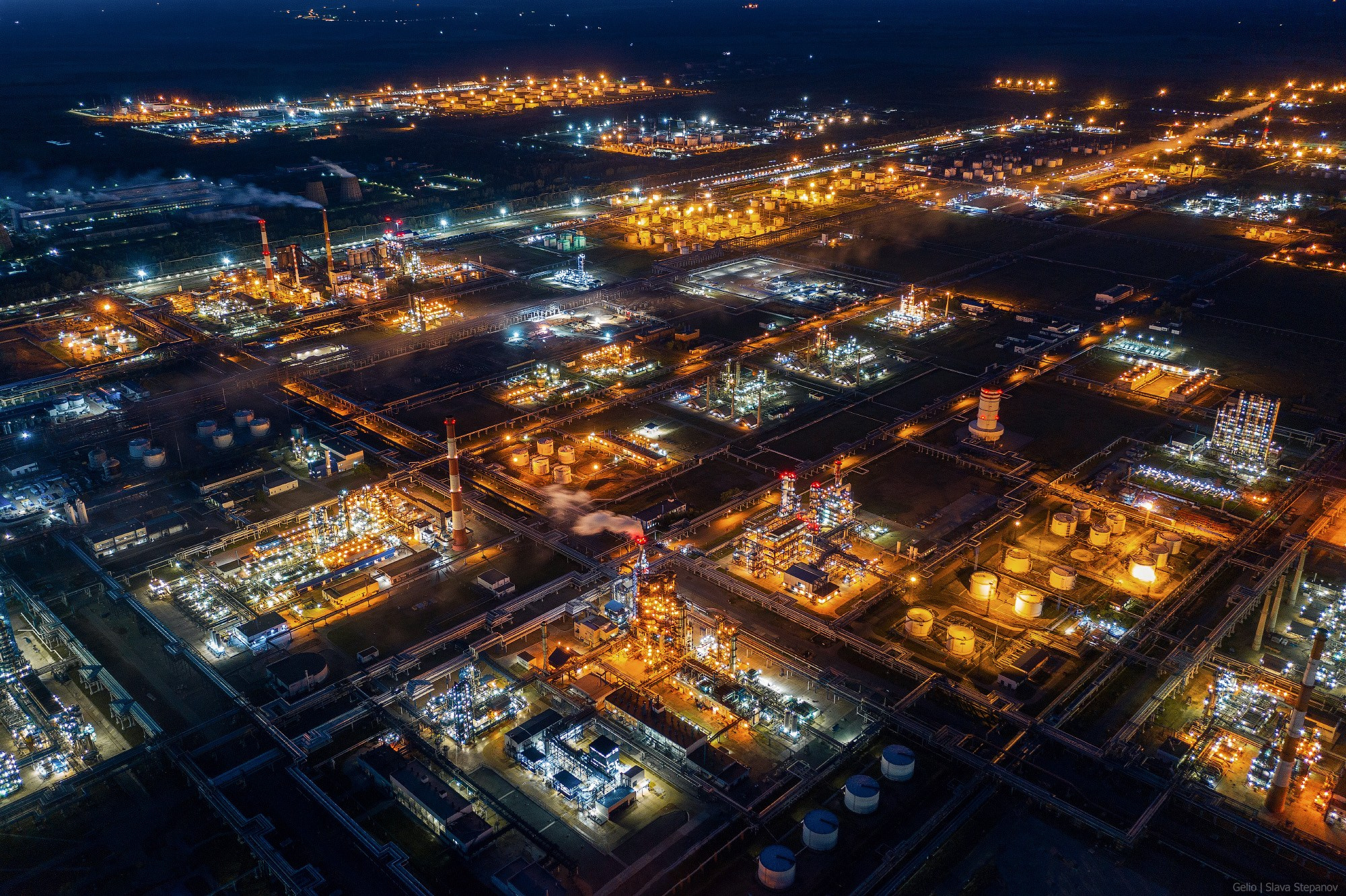 Огни гигантского нефтезавода в сумерках, плотная промышленная застройка и чёткая геометрия ярко освещённых технологических установок с высоты птичьего полёта, фото Слава Степанов.