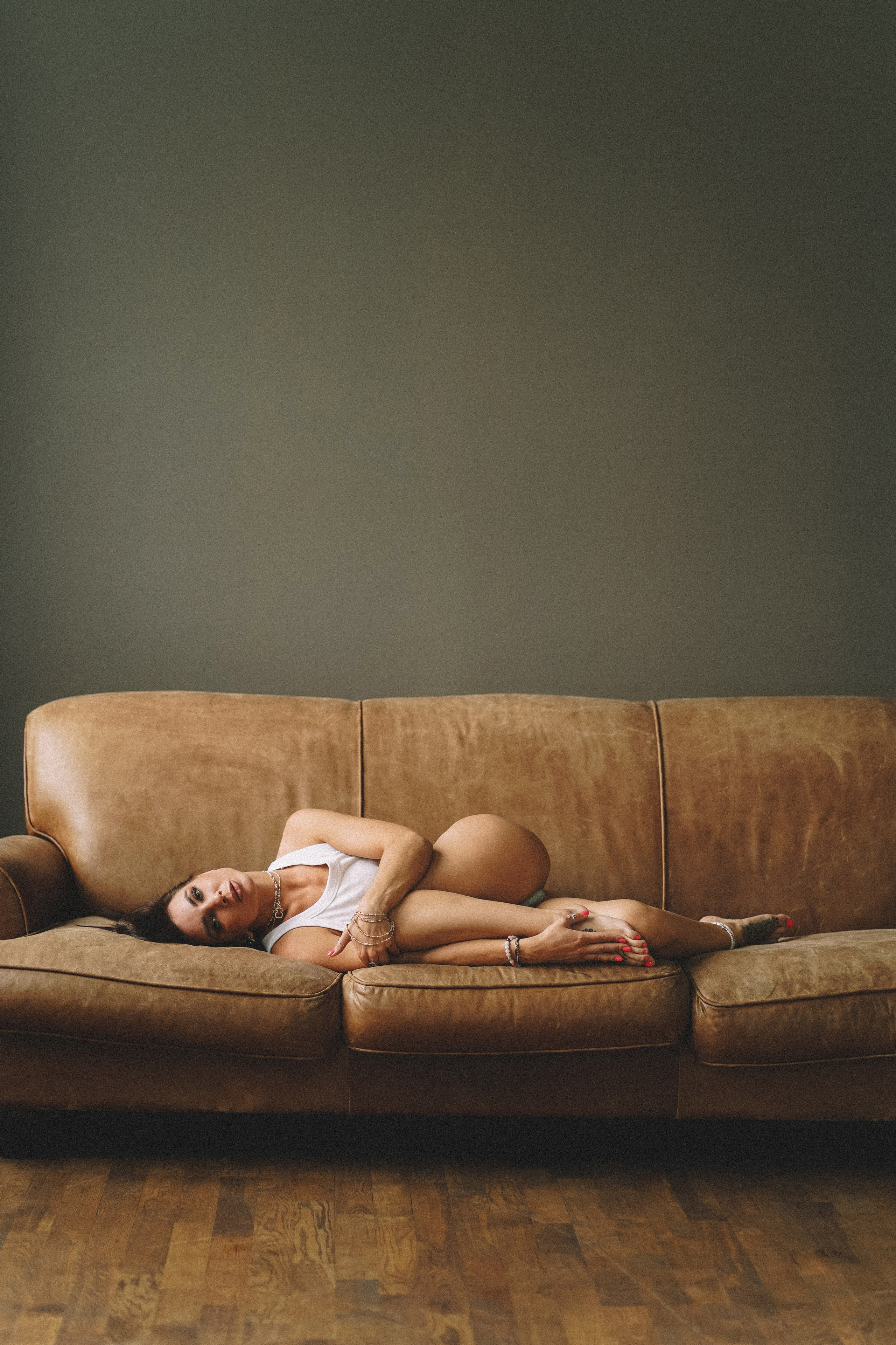 Девушка с каштановыми волосами в белой майке лежит на кожаном коричневом диване 