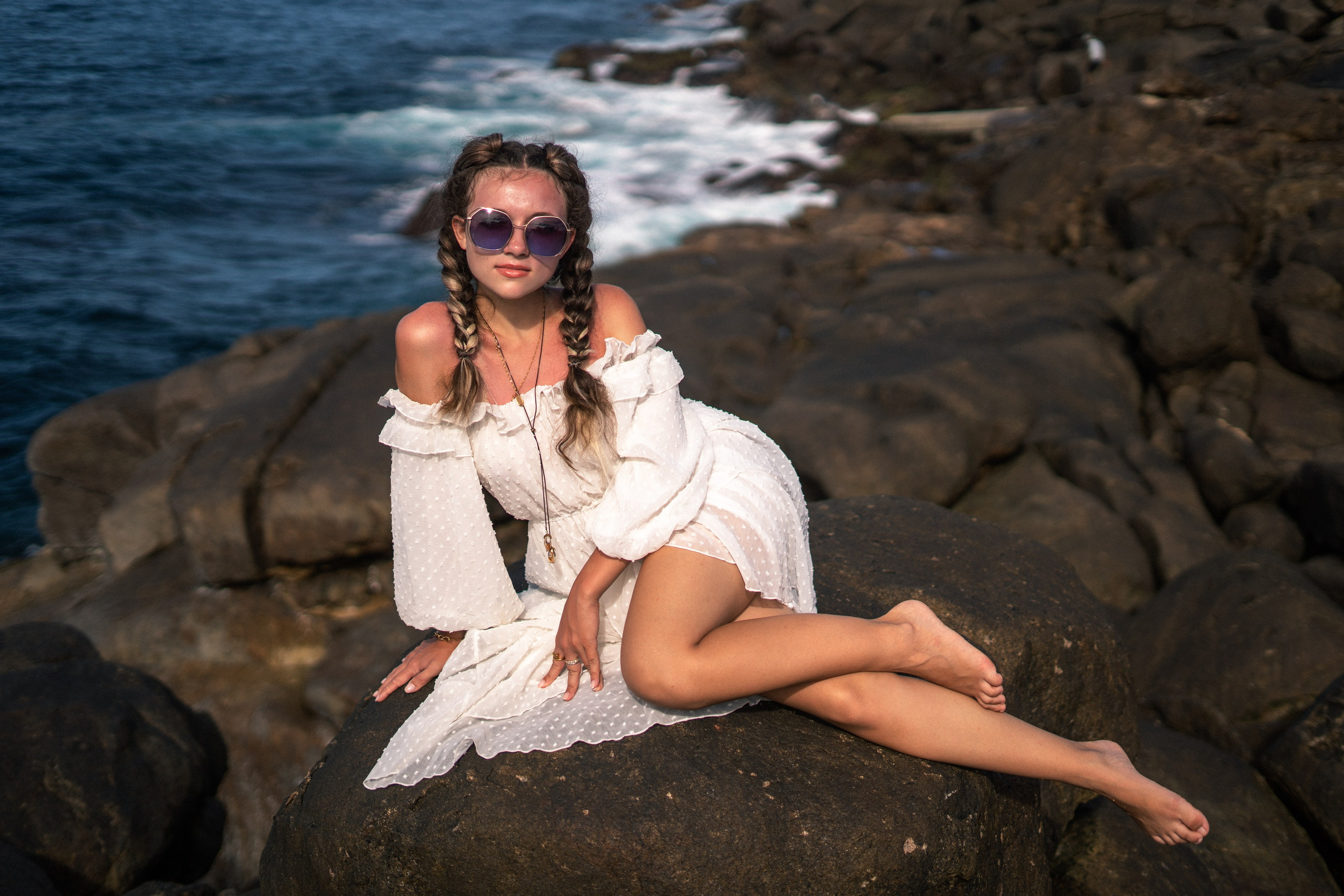 a girl in a white dress and glasses standing at the water's edge on the rocks