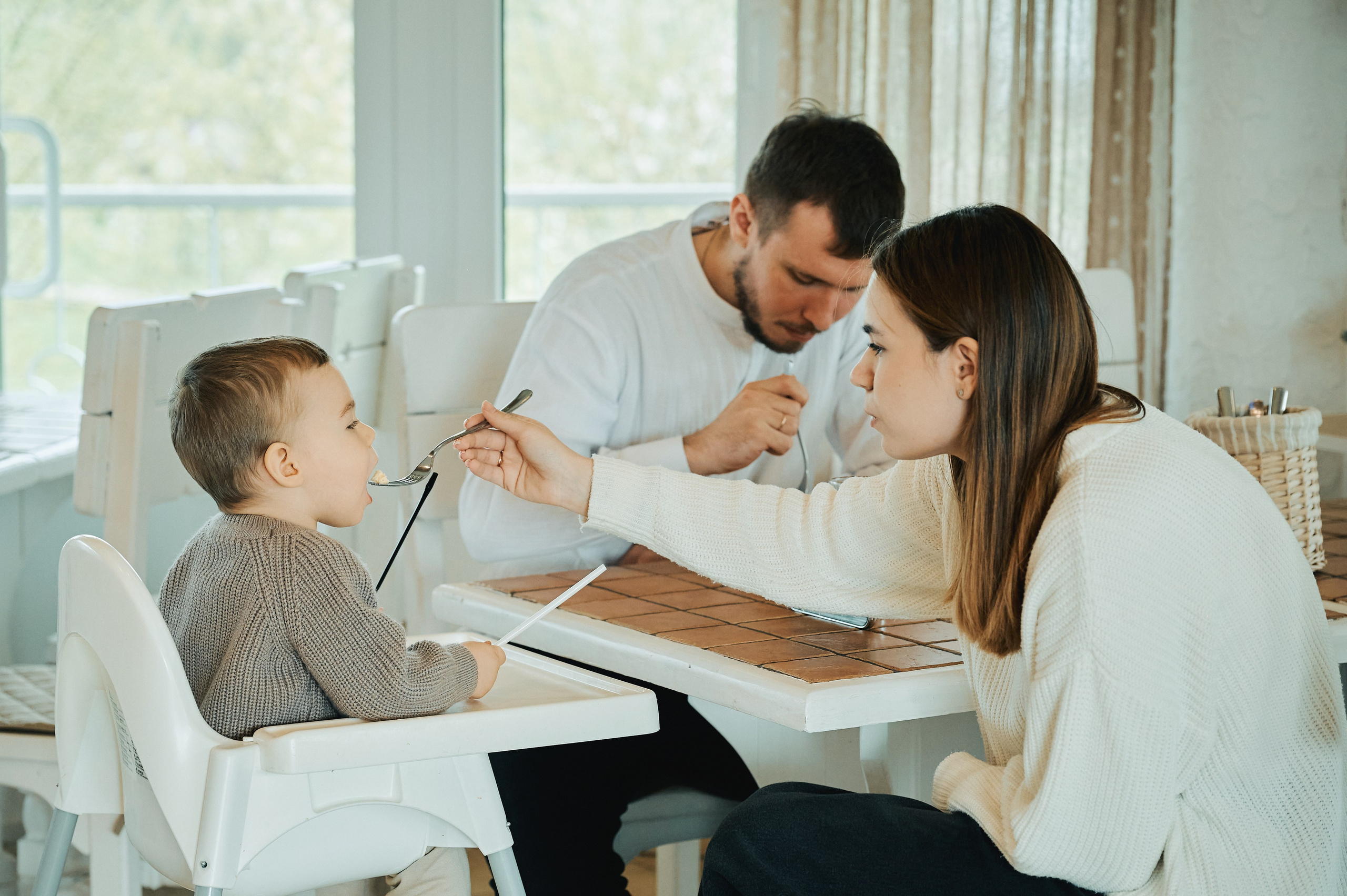 Мама кормит маленького сына с ложки за столом в светлой столовой, папа ест рядом.
