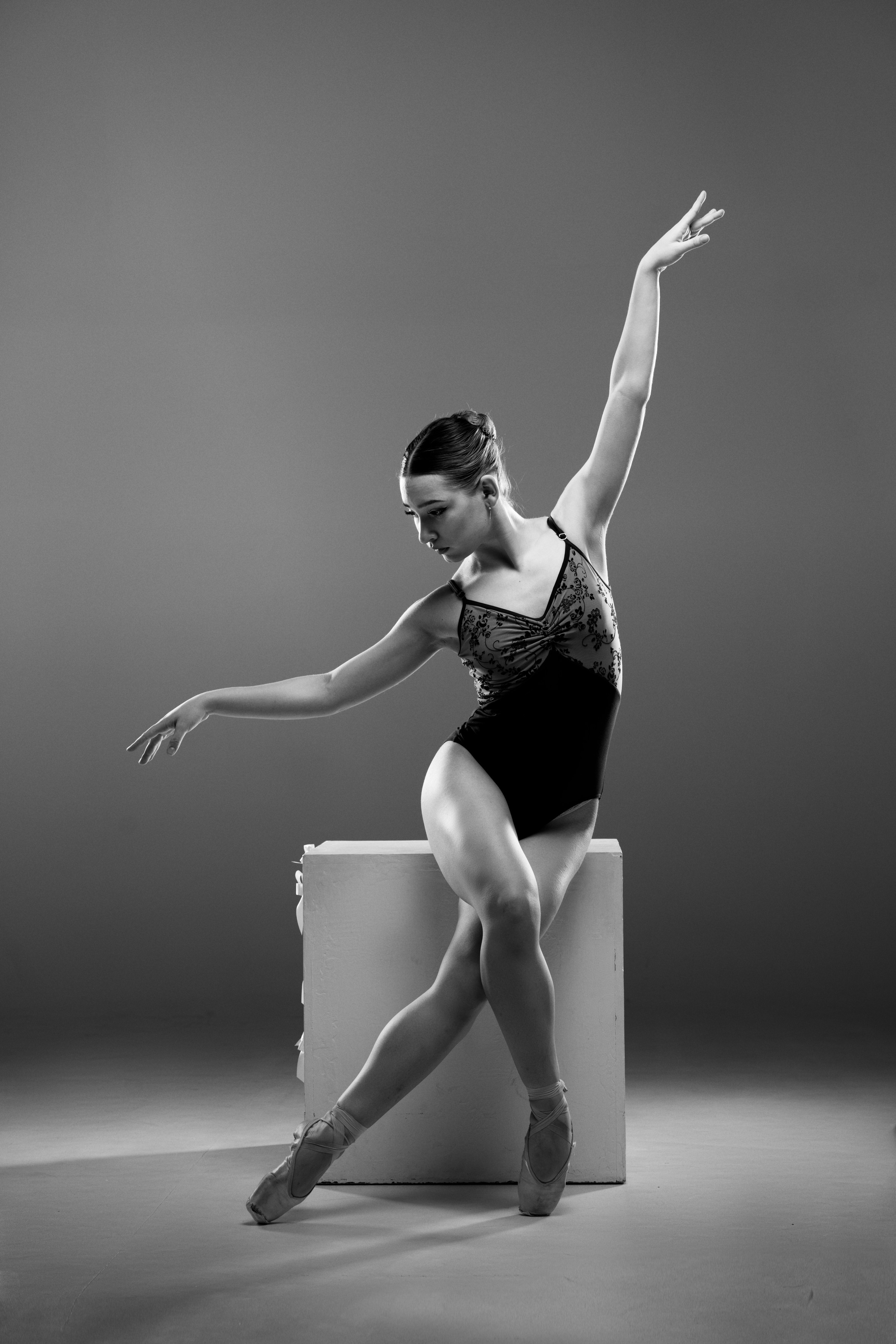 Спортивный фотограф в Тюмени: художественная балетная съемка в стиле спорт-арт. Черно-белый кадр балерины в черном купальнике, изящно позирующей на белом кубе. Акцент на элегантности жеста, чистоте линий и профессиональной балетной технике.