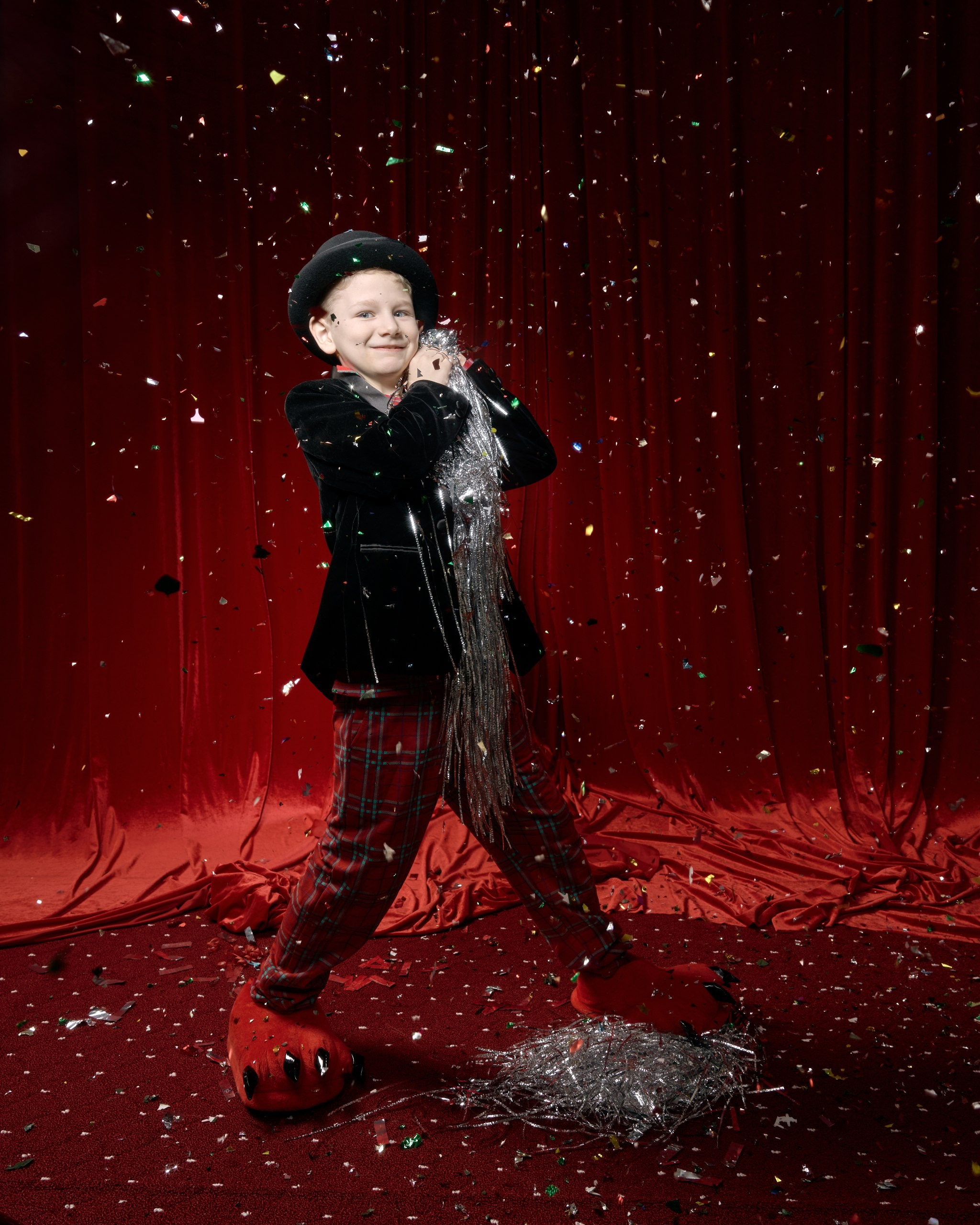 Фотосьемка с хлопушкой в фотостудии, разноцветная фольга. Яркая студийная съемка для детей: мальчик в костюме актера с серебряной гирляндой на фоне красного занавеса среди летящего конфетти. Семейный фотограф создает праздничные воспоминания.