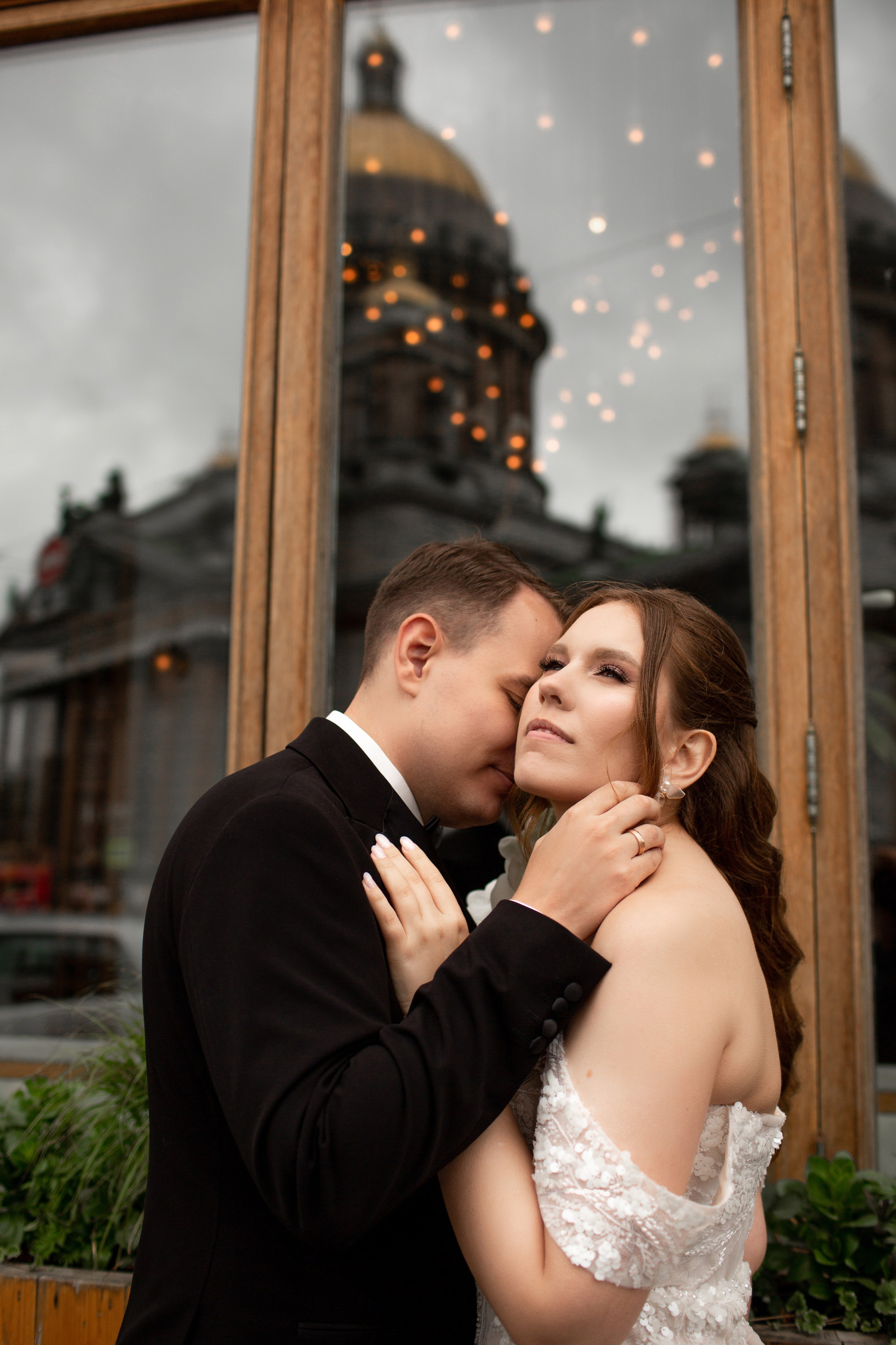 Красивая фотография: жених целует невесту в щёку, за ними в стекле отражается Исаакиевский собор.