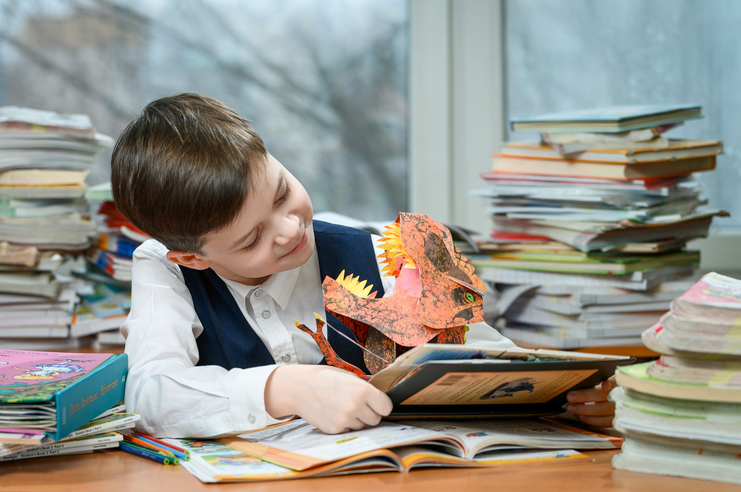 Мальчик рассматривает книгу за партой среди стопок учебников