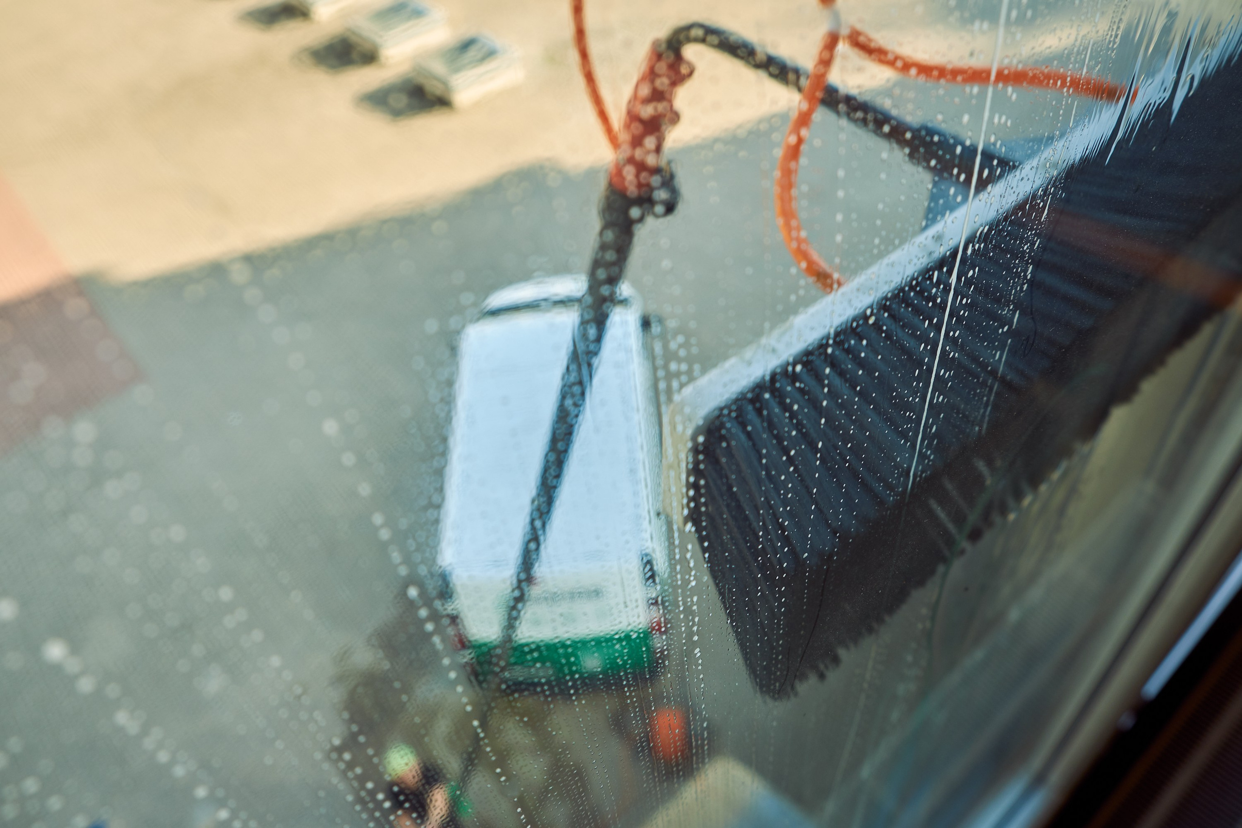 Фотография процесса мойки окон снаружи здания, снятая изнутри помещения. Крупный план инструмента и капель воды. Коммерческая съёмка рабочих процессов для клининговых услуг.