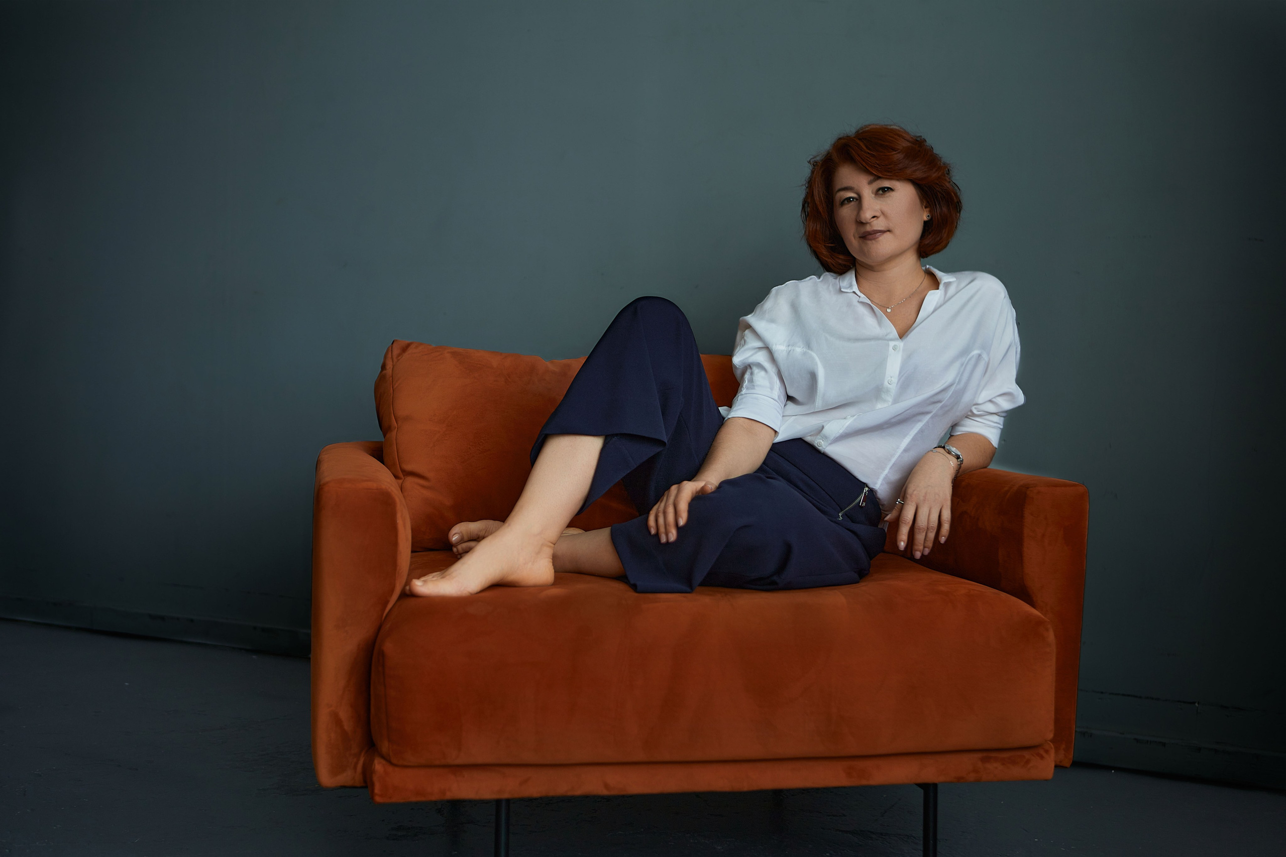 Женщина в белой рубашке и брюках сидит в кресле терракотового цвета