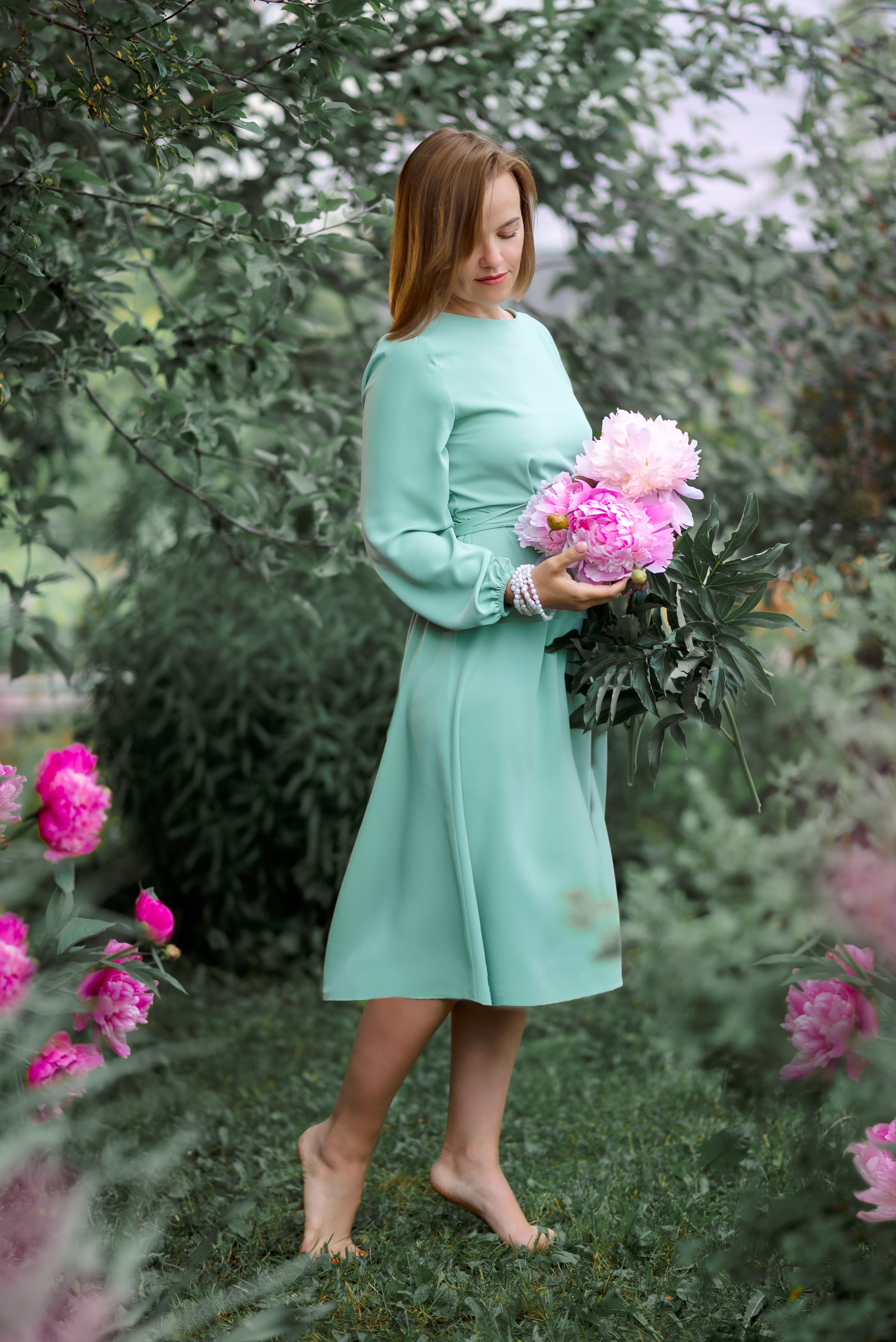 Девушка в полный рост с букетом пионов стоит в саду в зеленом платье