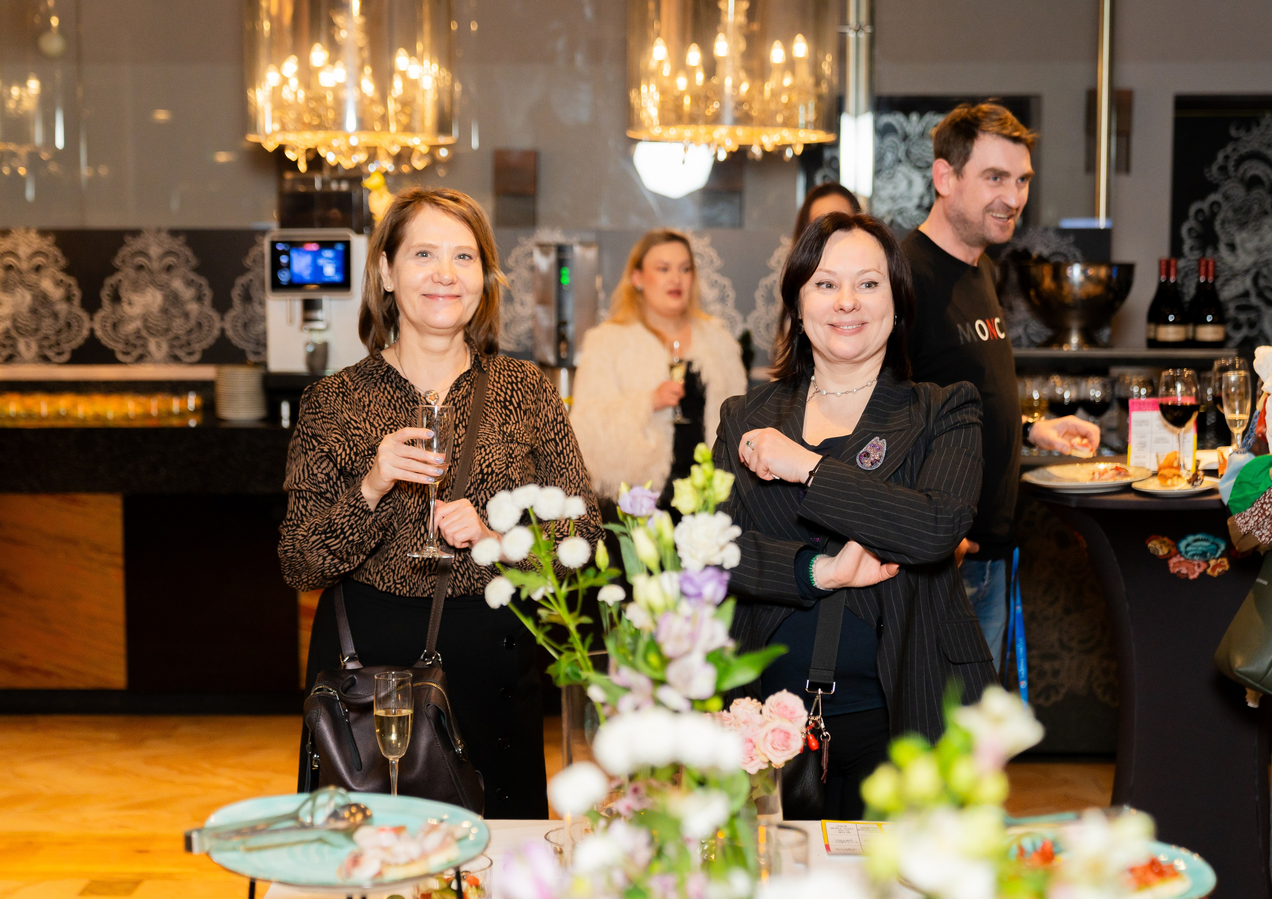 Гости вечеринки в Radisson Sonya: женщина в леопардовой блузке и женщина в полосатом пиджаке с бокалами шампанского. На заднем плане мужчина в футболке Moncler. Фото Евгения Мальцева.