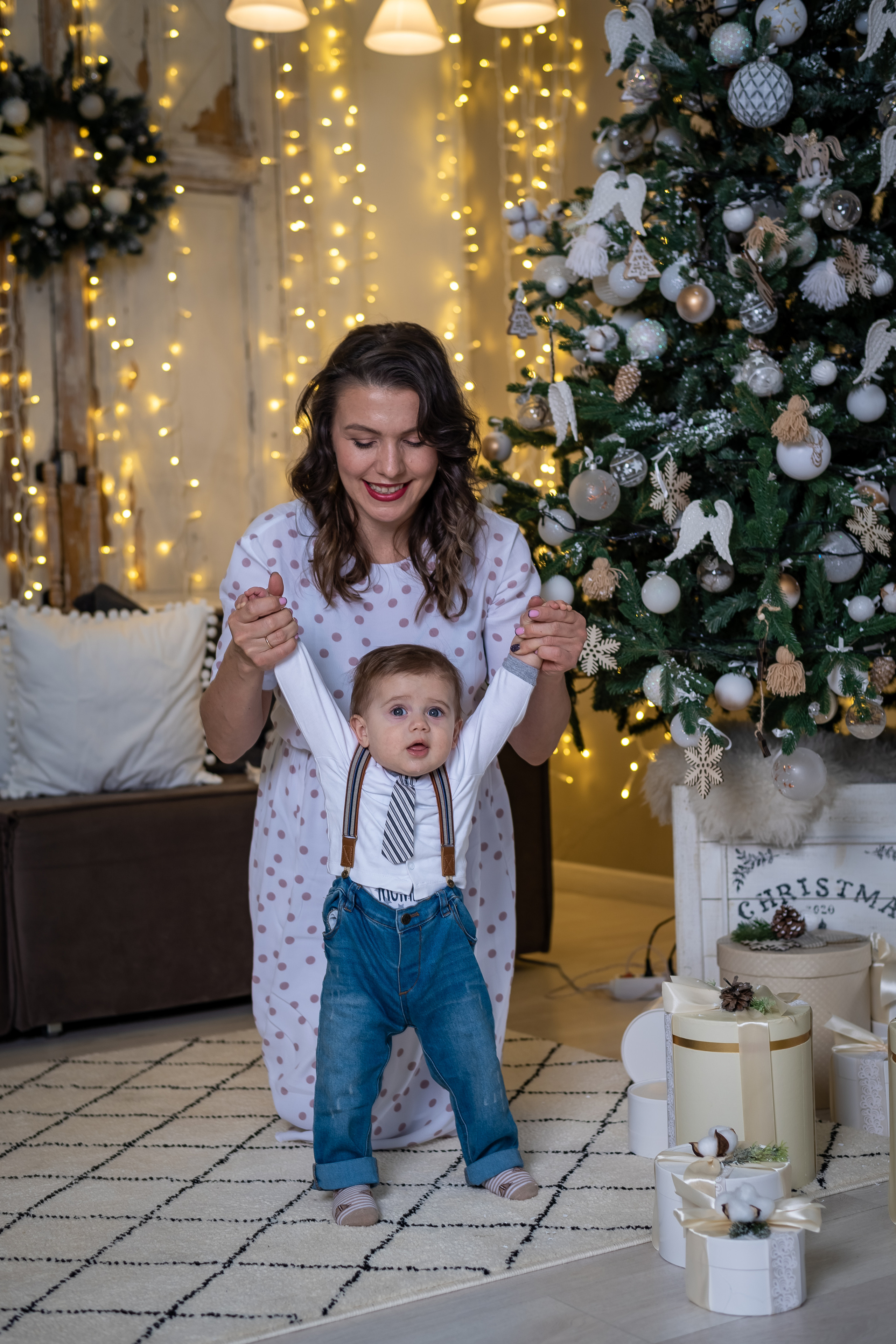 Мама держит за ручки малыша на фоне украшенной елки.