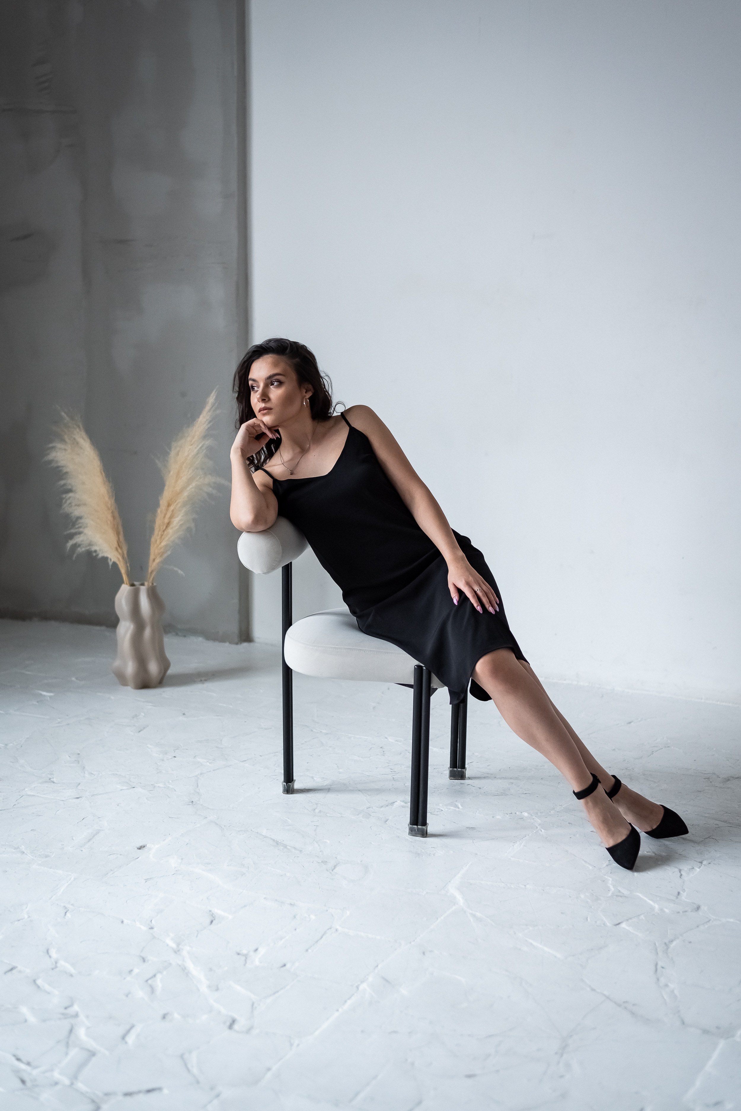 девушка сидящая на стуле в черном платье с вазой на заднем плане на фоне белая и серая стена