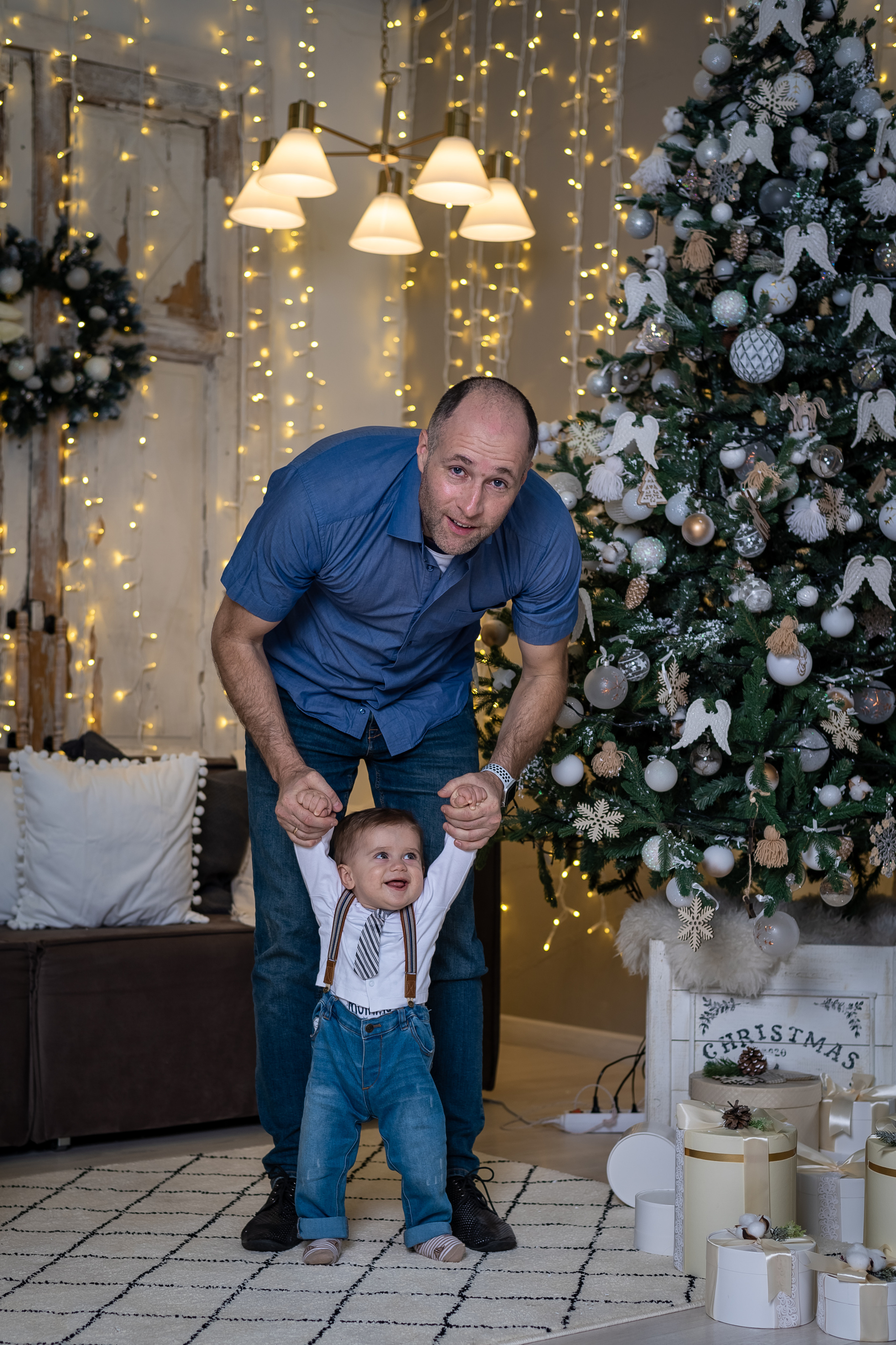 папа держит за ручки малыша возле новогодней елки