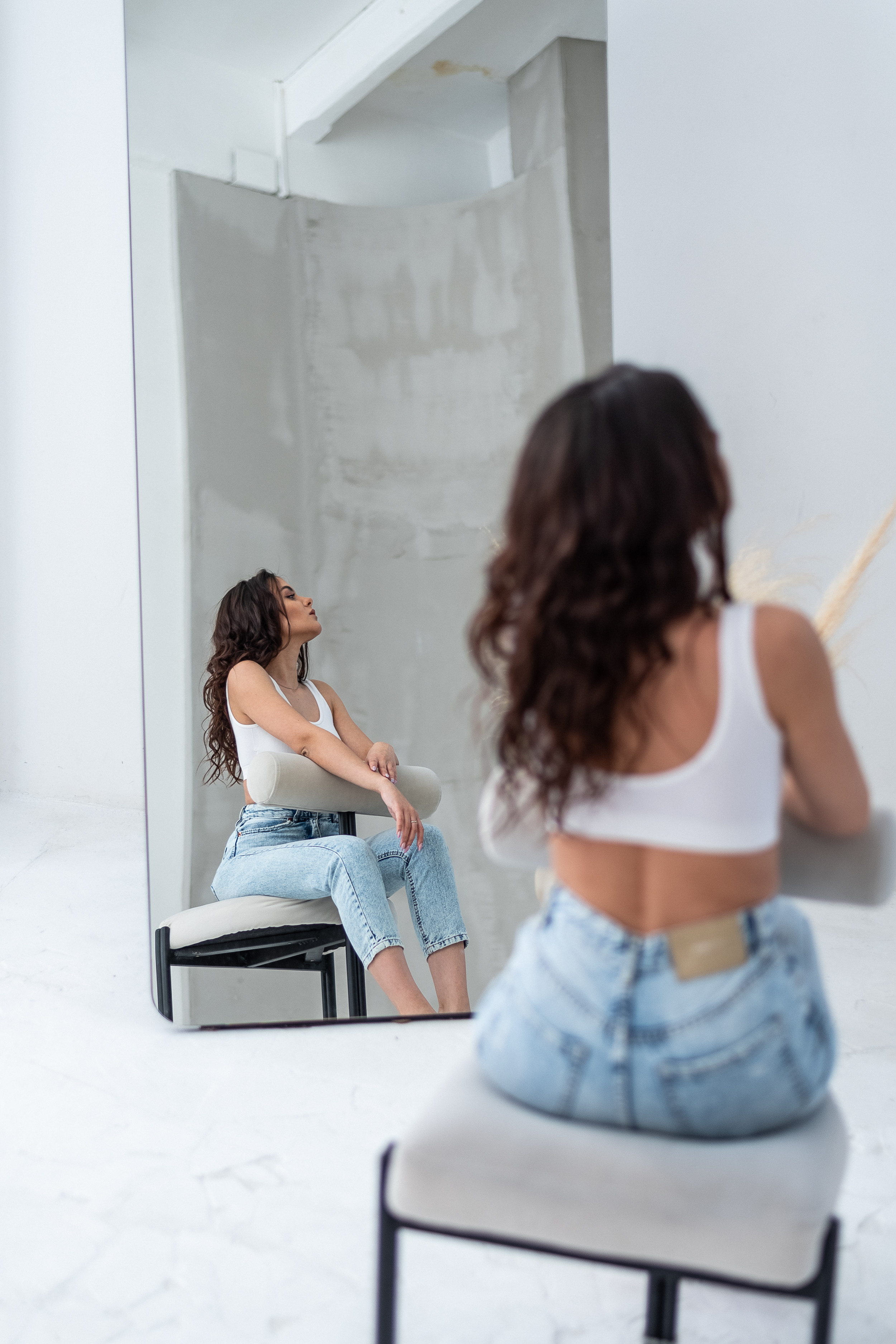 отражение девушки с темными волосами ,сидящей на стуле, в голубых джинсках 