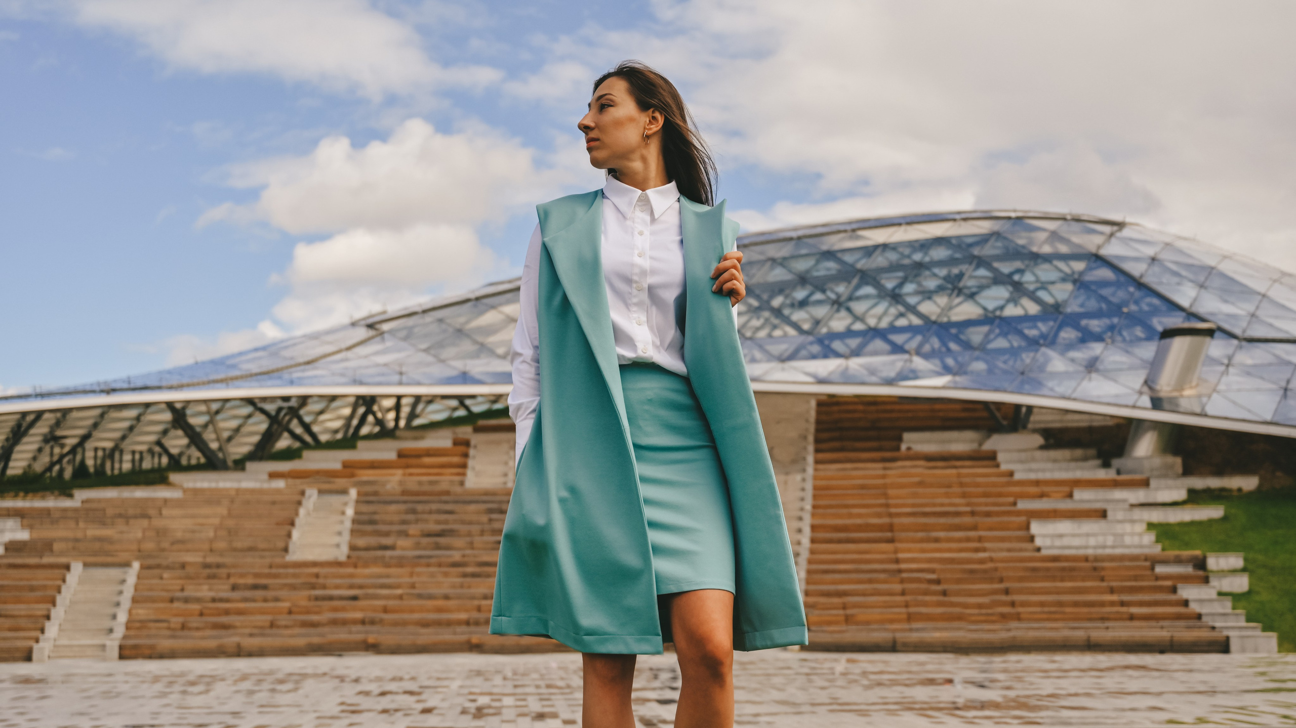 Молодая женщина в элегантном образе с белой блузкой, светло-зеленой юбкой и жакетом стоит на фоне современной архитектуры. Она одета в стильный комплект, который идеально сочетается с окружающей средой. Волосы аккуратно собраны назад, а ее взгляд направлен в сторону, создавая ощущение уверенности и задумчивости. На заднем плане видна структура здания с прозрачной крышей и деревянными лестницами, что добавляет фотографии элементы модернизма и контраст между человеком и городской средой. Светлый фон неба с легкими облаками подчеркивает свежесть и чистоту момента. Этот снимок демонстрирует профессиональное владение техникой освещения и композиции, делая акцент на игре света и тени, а также на гармоничном сочетании человека и современного окружения. Perfect for fashion and architectural photography enthusiasts!