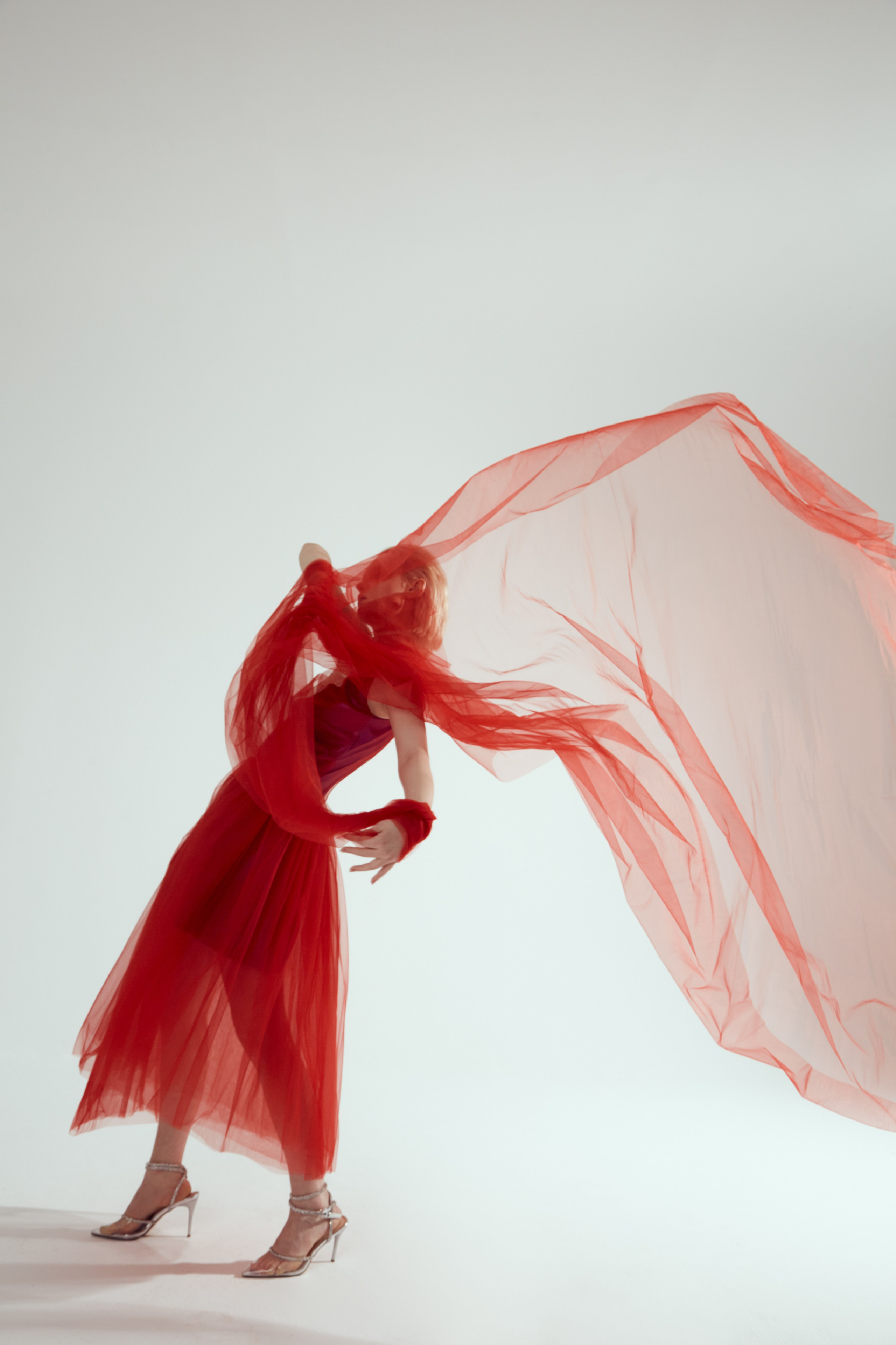 Девушка в легком красном платье и прозрачной ткани, развивающейся вокруг нее, стоит в динамичной позе на светлом фоне
