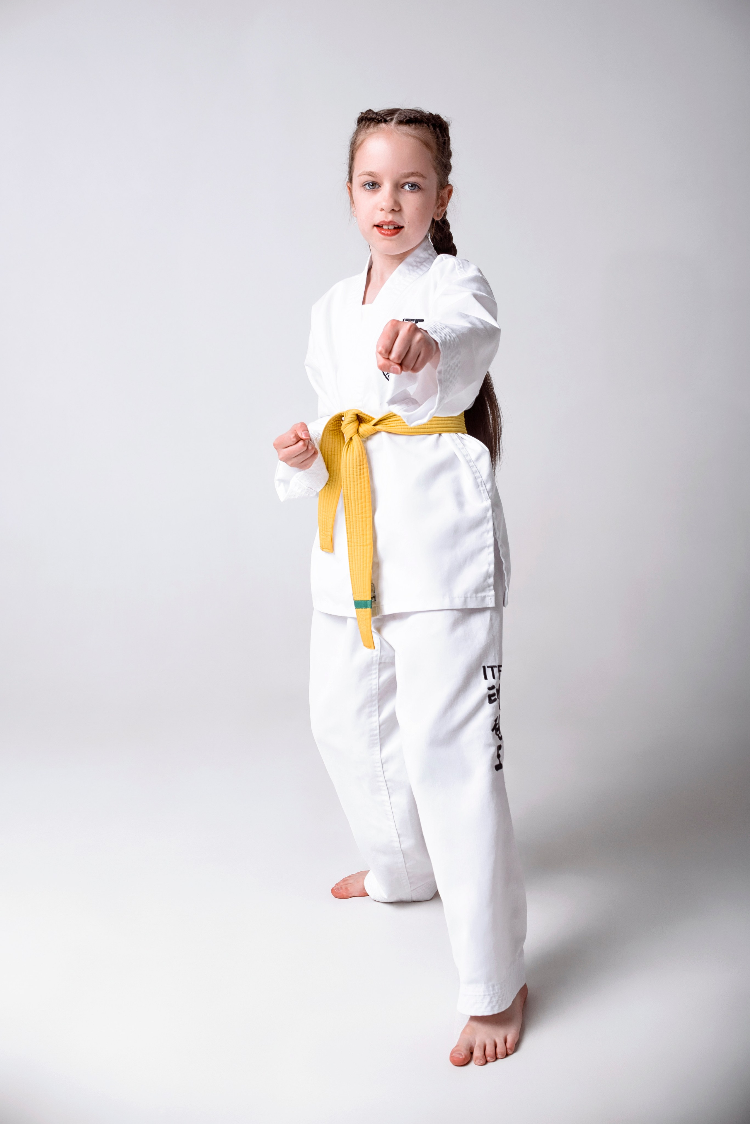 На изображении изображено лицо, одетое в белую форму Тхэквондо (добок) с желтым поясом. Человек носит красные боксерские перчатки и, вероятно, готовится к тренировке боевых искусств. На форме есть нашивки, одна из которых содержит буквы "ITF", что, вероятно, указывает на Международную федерацию Тхэквондо. Фон светлый и однородный, что обеспечивает четкий обзор объекта.