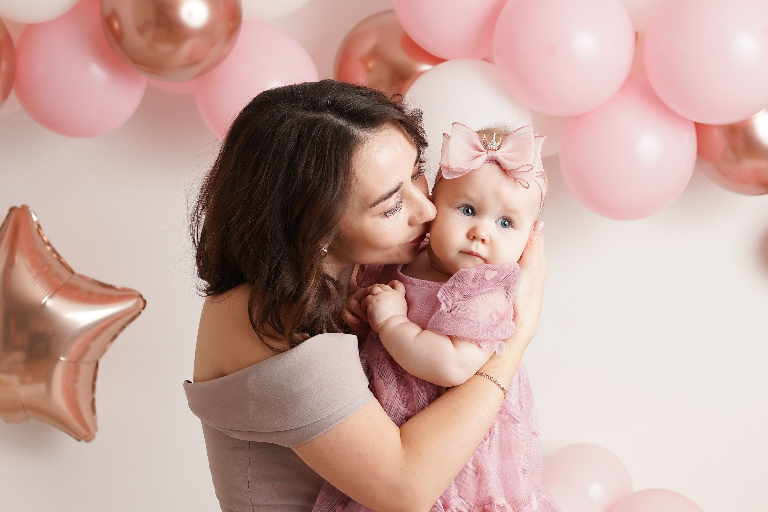 На фото девочка в красивом розовом платье. Ее держит на руках мама. Она ее нежно целует.