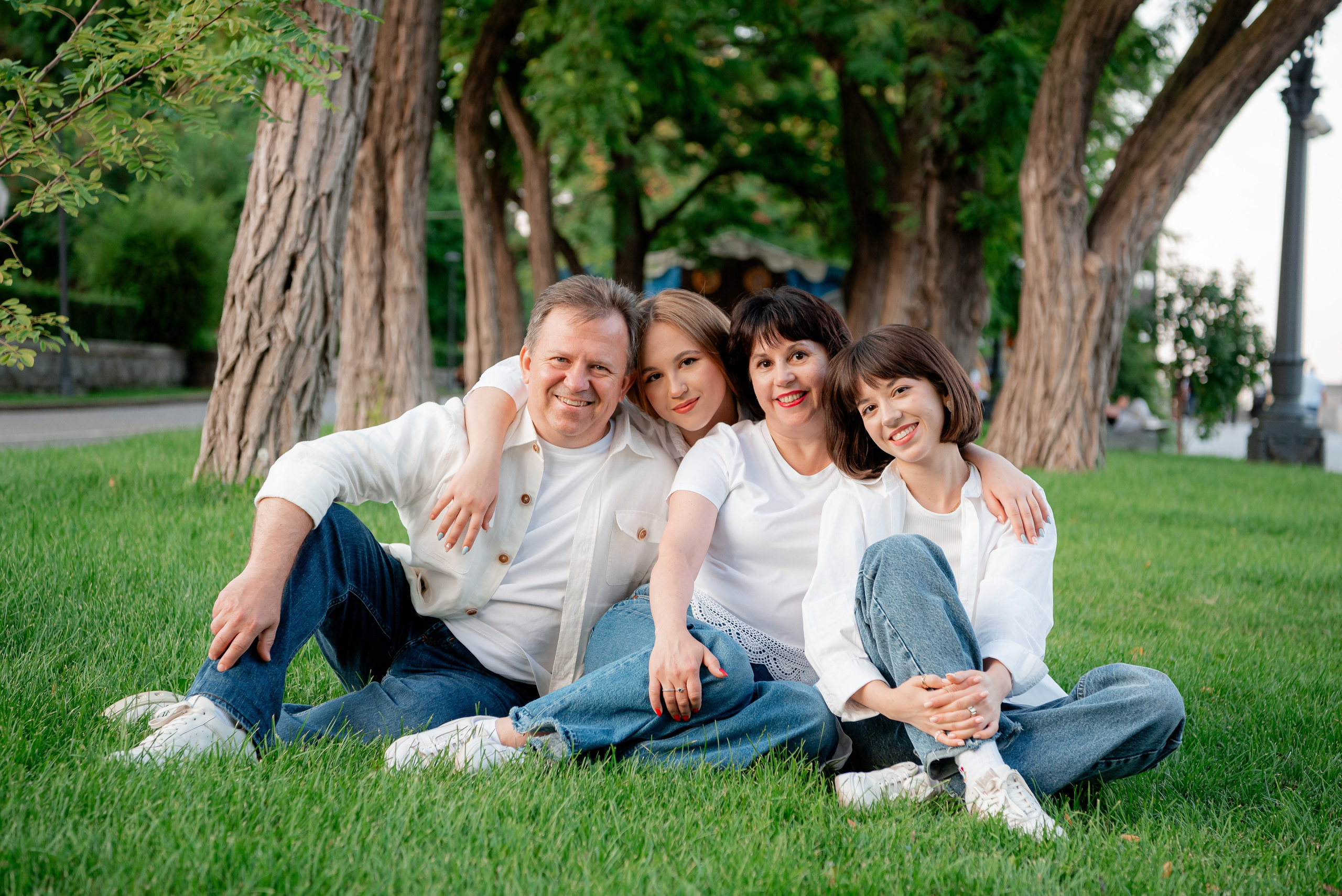 Постановочно семейный портрет сидя на траве большой семьи папа мама и две взрослых дочки все одеты белый верх и синие джинсы