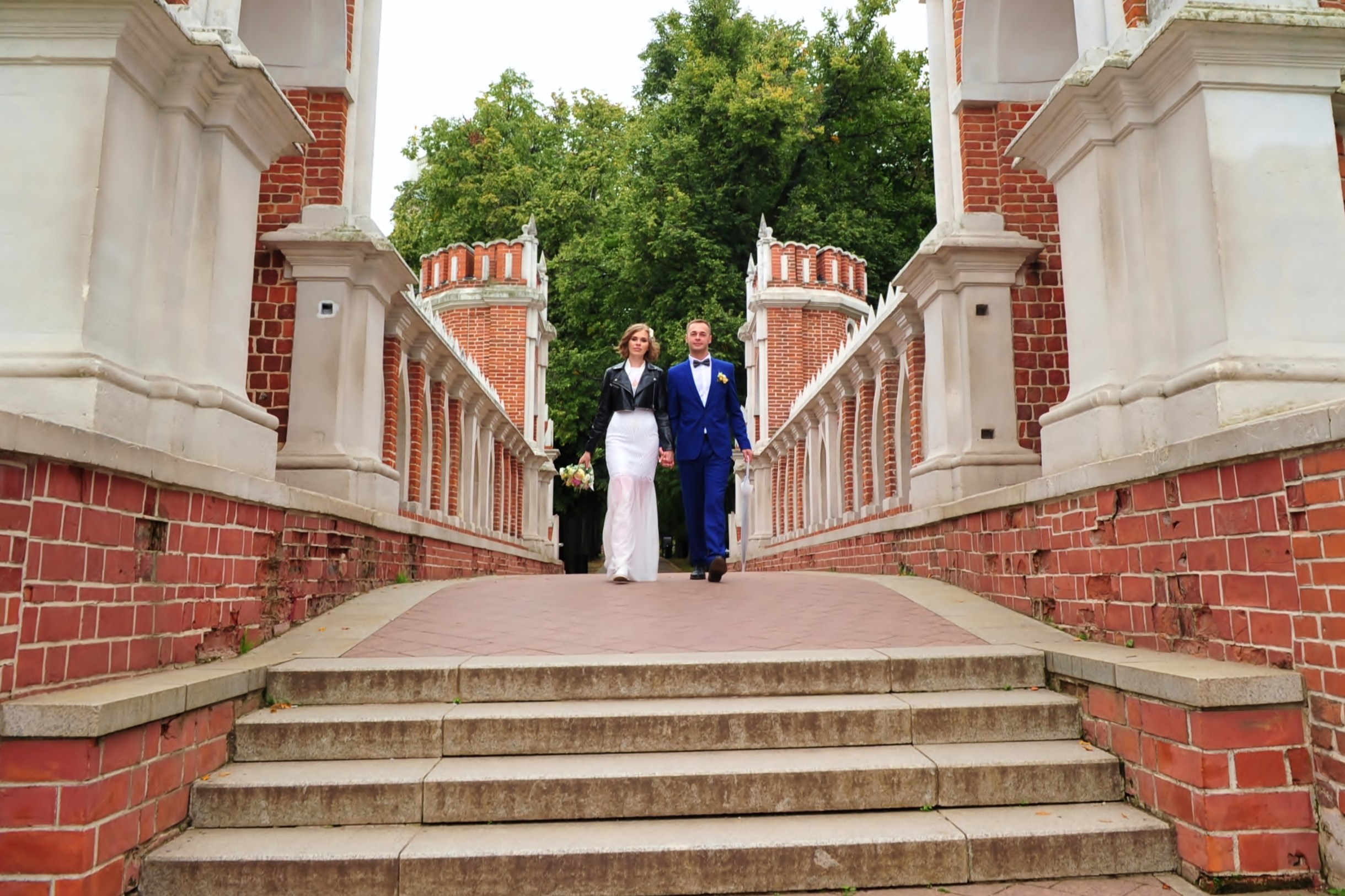 Свадьба в Царицыно хмурым прохладным летним днём, молодожены идут на мосту из красного кирпича, держатся за руки и смотрят вперед