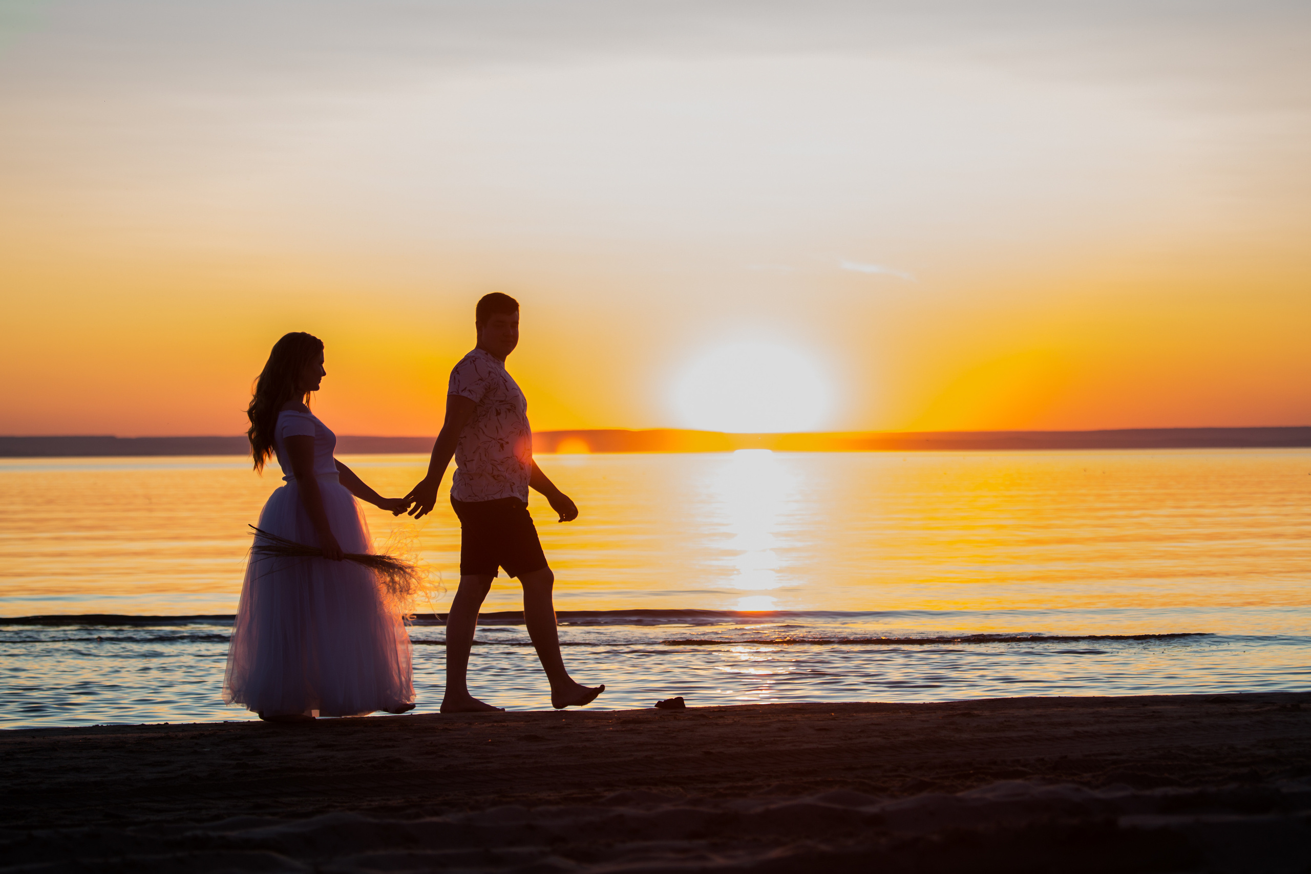 молодая пара идет по берегу на фоне моря и заката