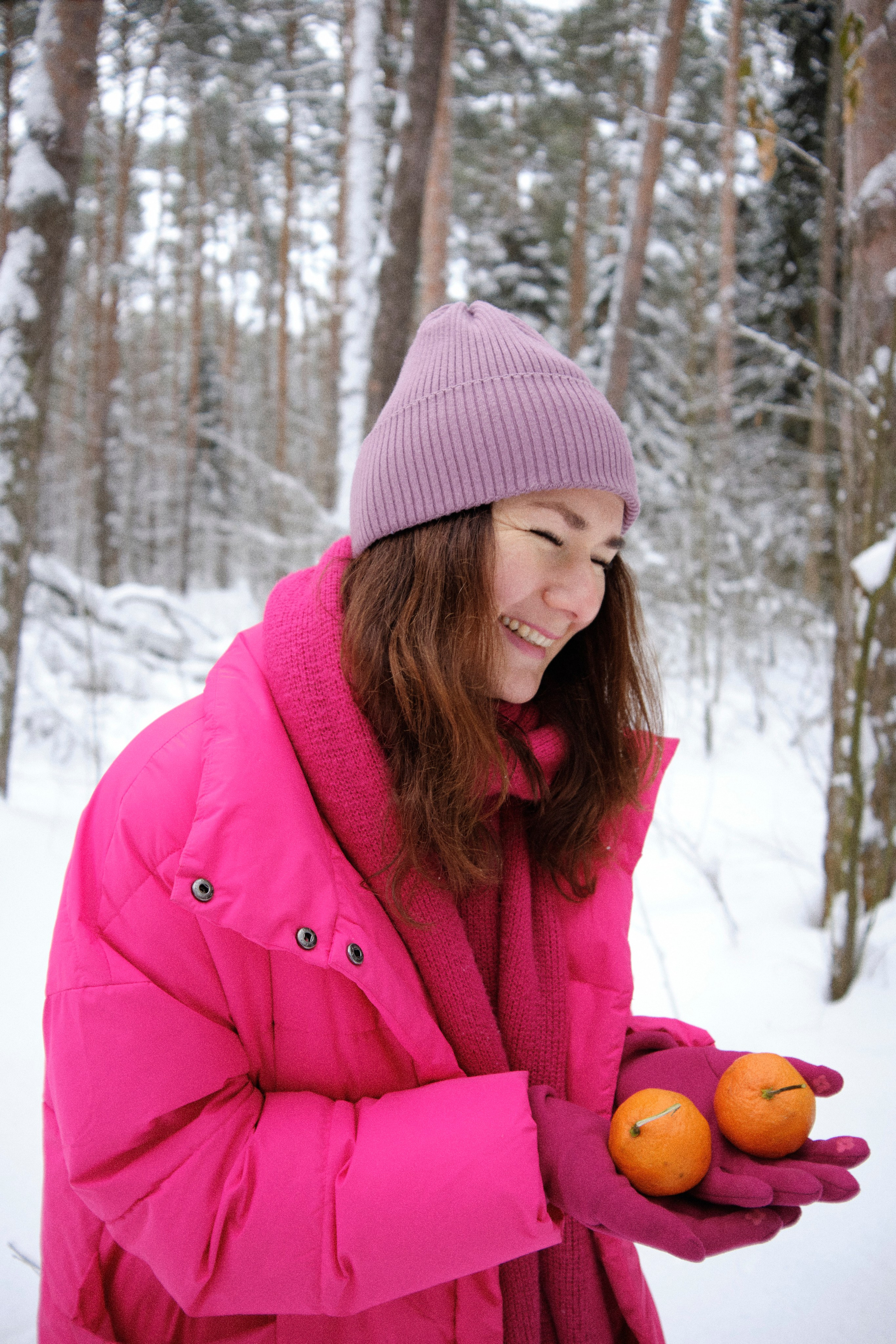 Женщина в яркой одежде смеется в зимнем лесу с мандаринами в руках