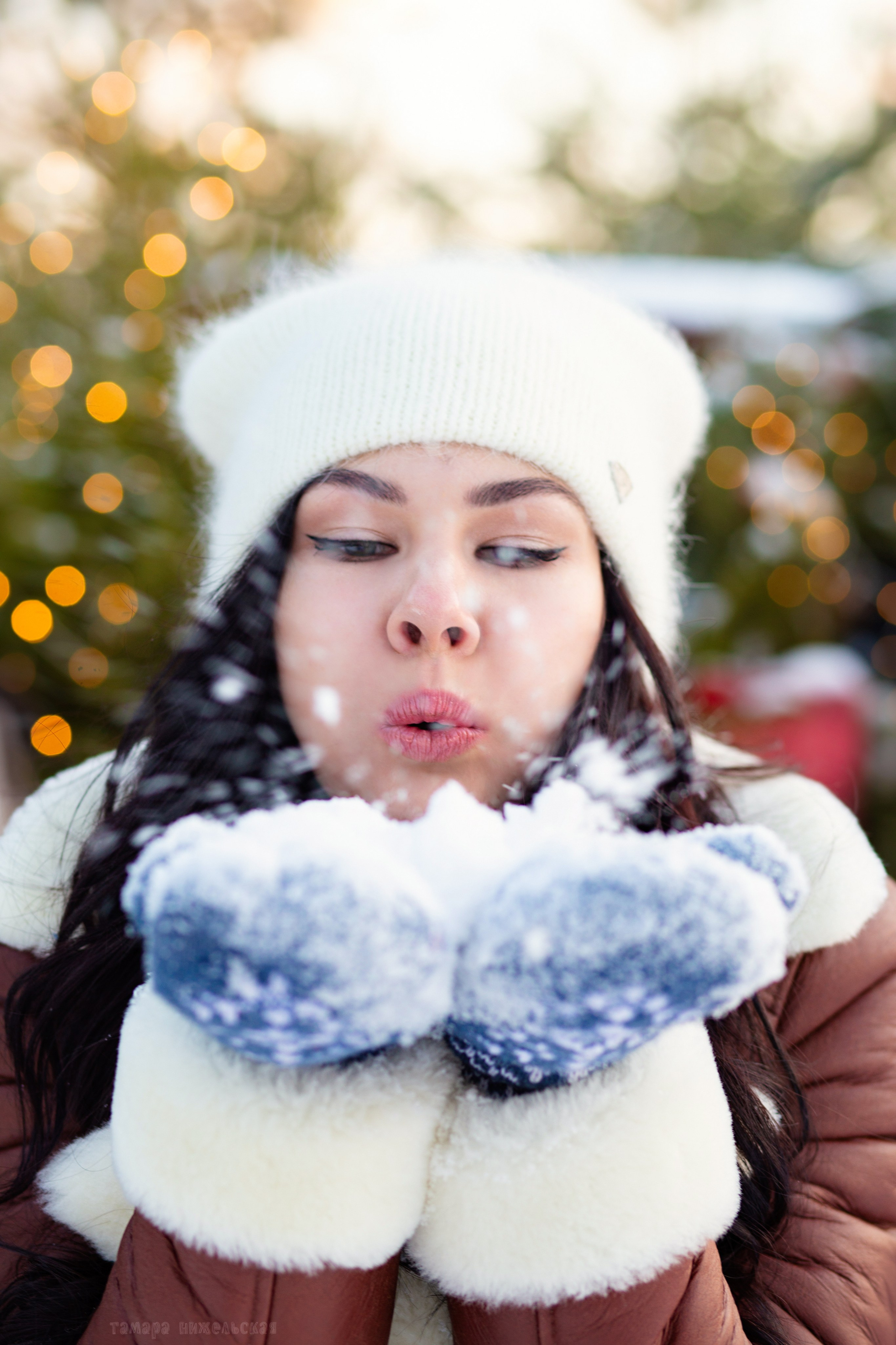 девушка в белой  шапке, коричневой   куртке и синих варежках  держит руки у лица. в открытых ладонях снег. она  дует на  снег. снег  летит  вперёд. 