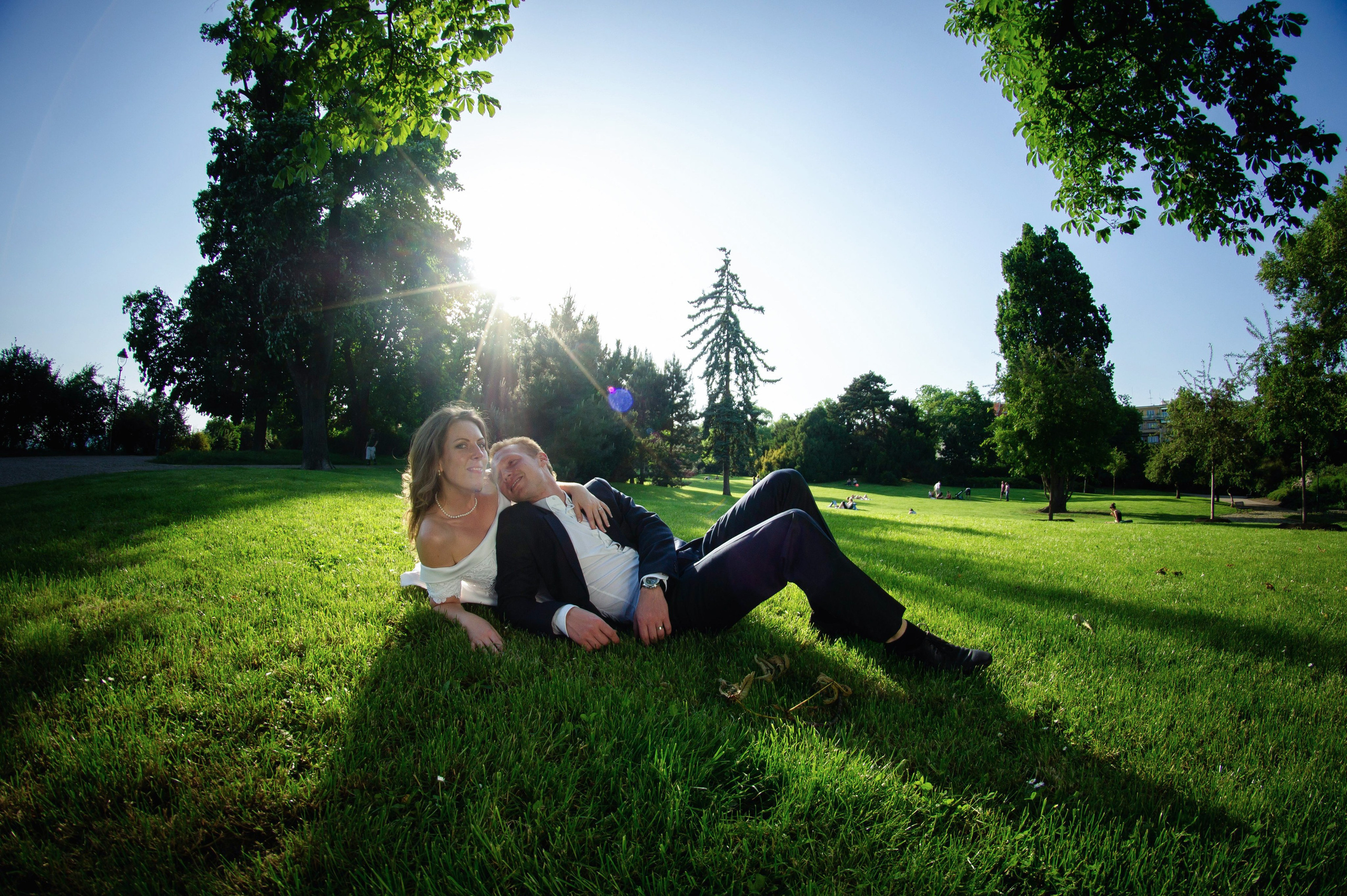 Молодожены лежат на лужайке среди высоких деревьев.