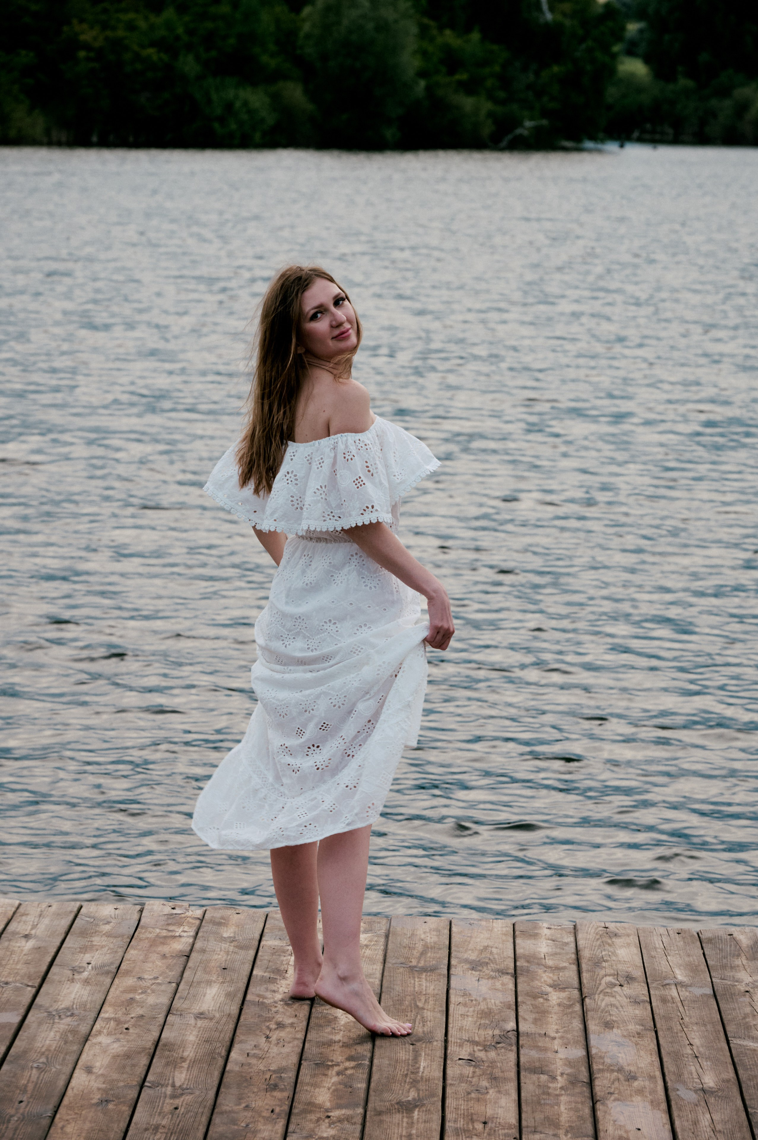 Евгения идёт босиком по деревянному пирсу у озера в белом платье.