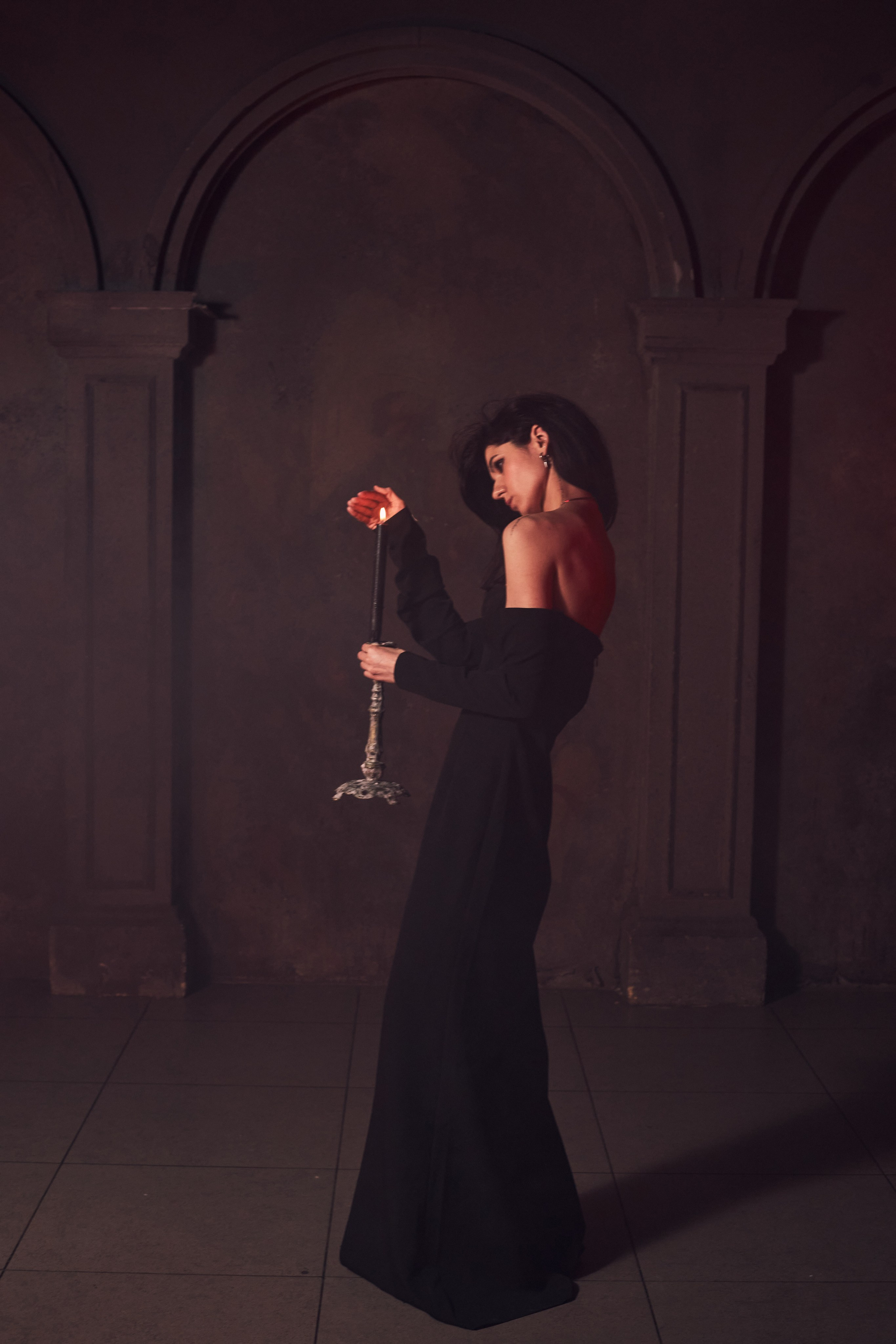 Девушка в длинном черном платье с открытыми плечами держит в руках свечу в подсвечнике