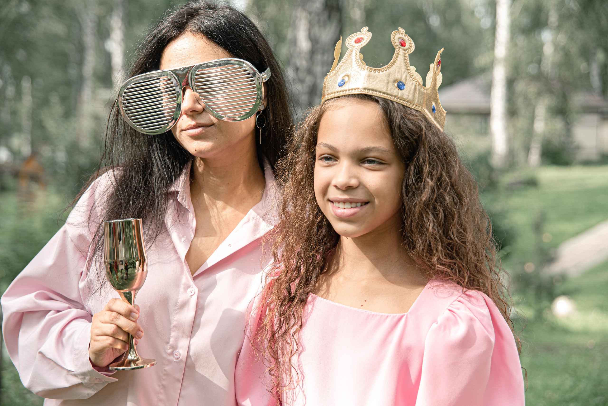 На фото женщина и дочка в розовых нарядах, одна из которых держит бокал с напитком. У одной из них на голове корона, что придает образу праздничный вид. Фон украшен зелеными деревьями и природой, создавая атмосферу веселья и торжества.
