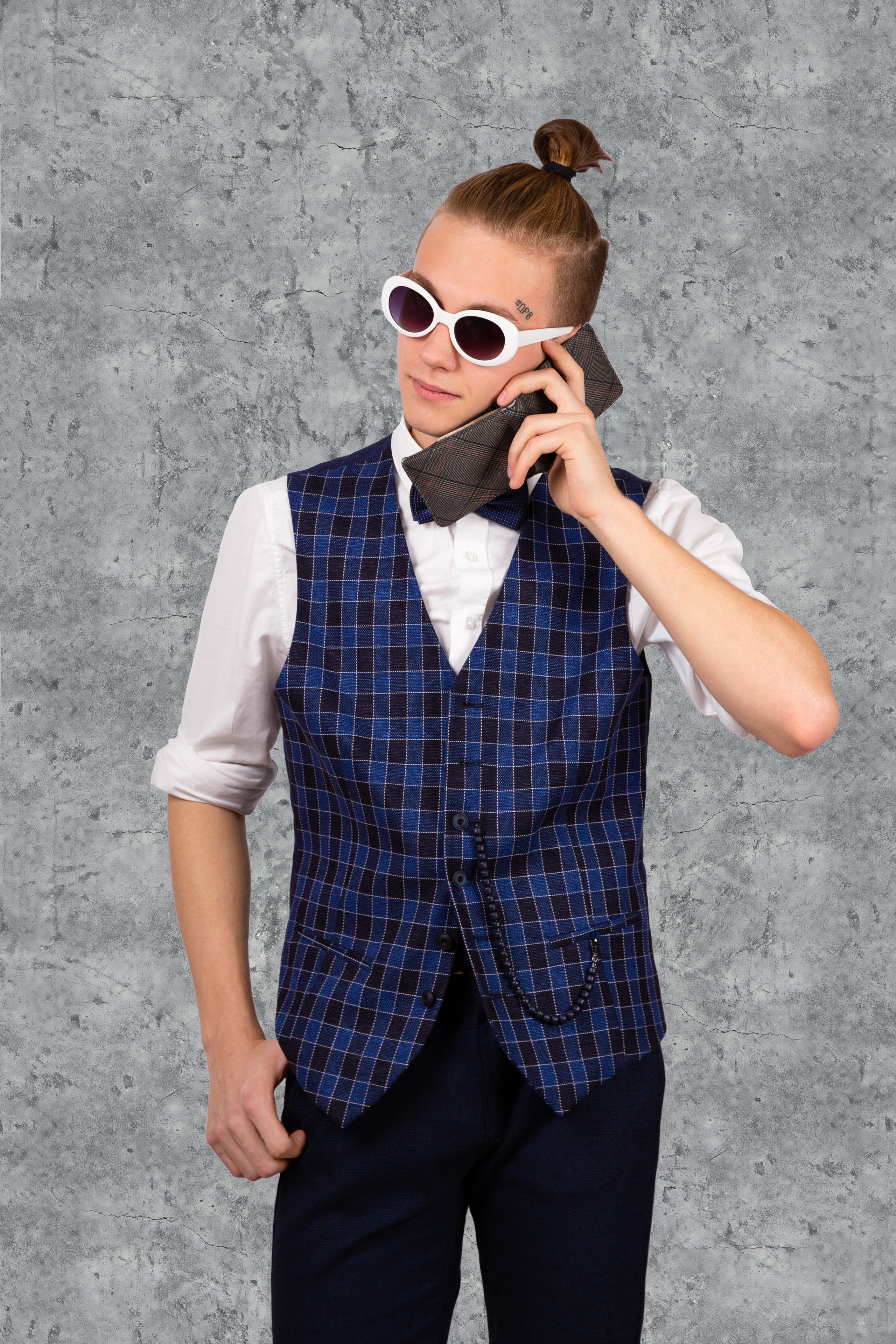 Юноша в жилетке, солнечных очках и с хвостиком делает вид, что разговаривает по бумажнику.