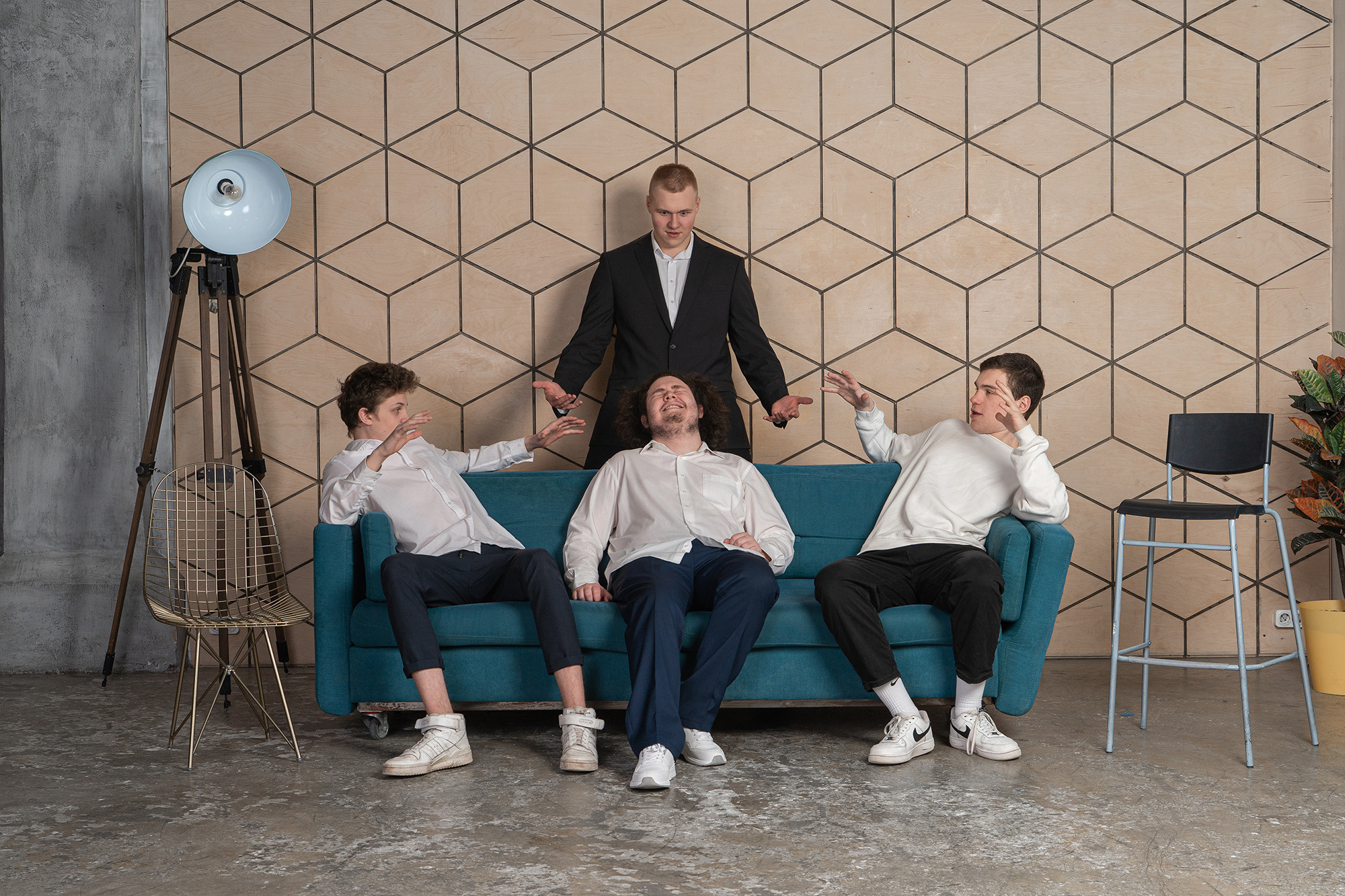 Трое юношей сидят на синем диване. Сзади дивана стоит юноша в пиджаке. Слева от дивана - прожектор.