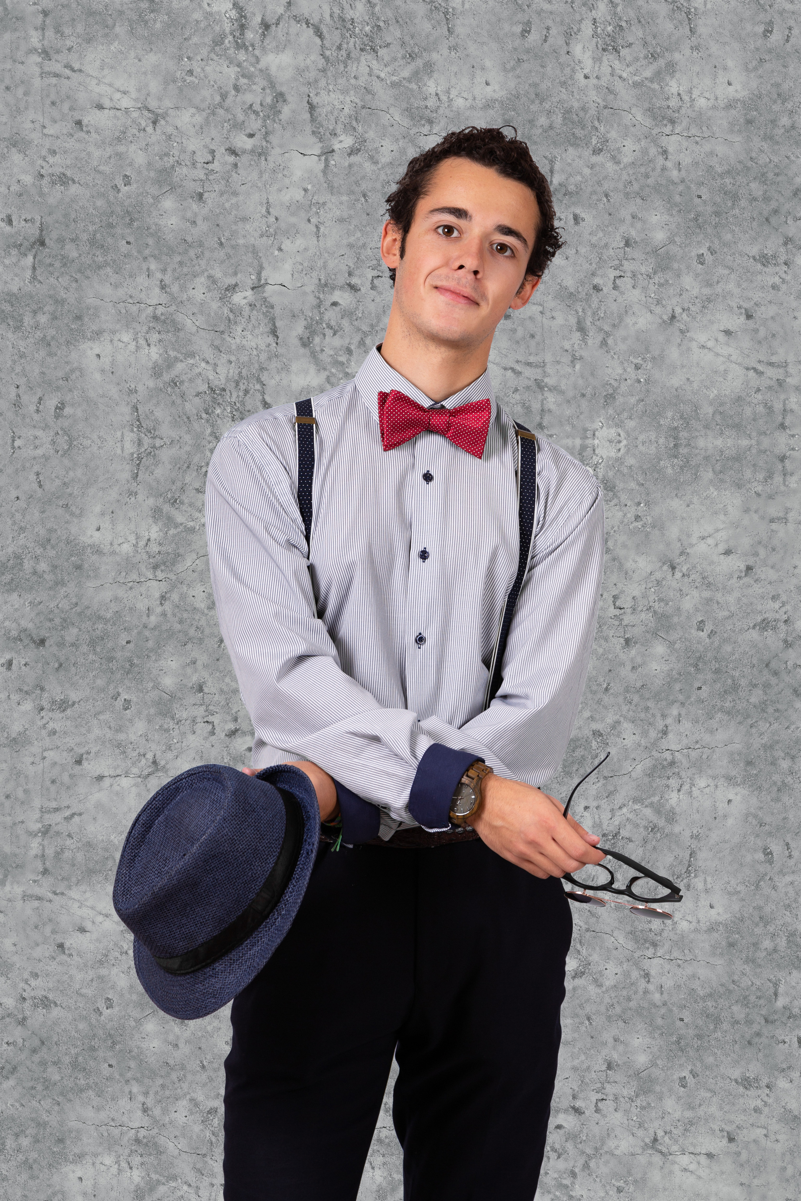 Портрет юноши для выпускного альбома. Юноша на фоне серой бетонной стены, в руках очки и шляпа.