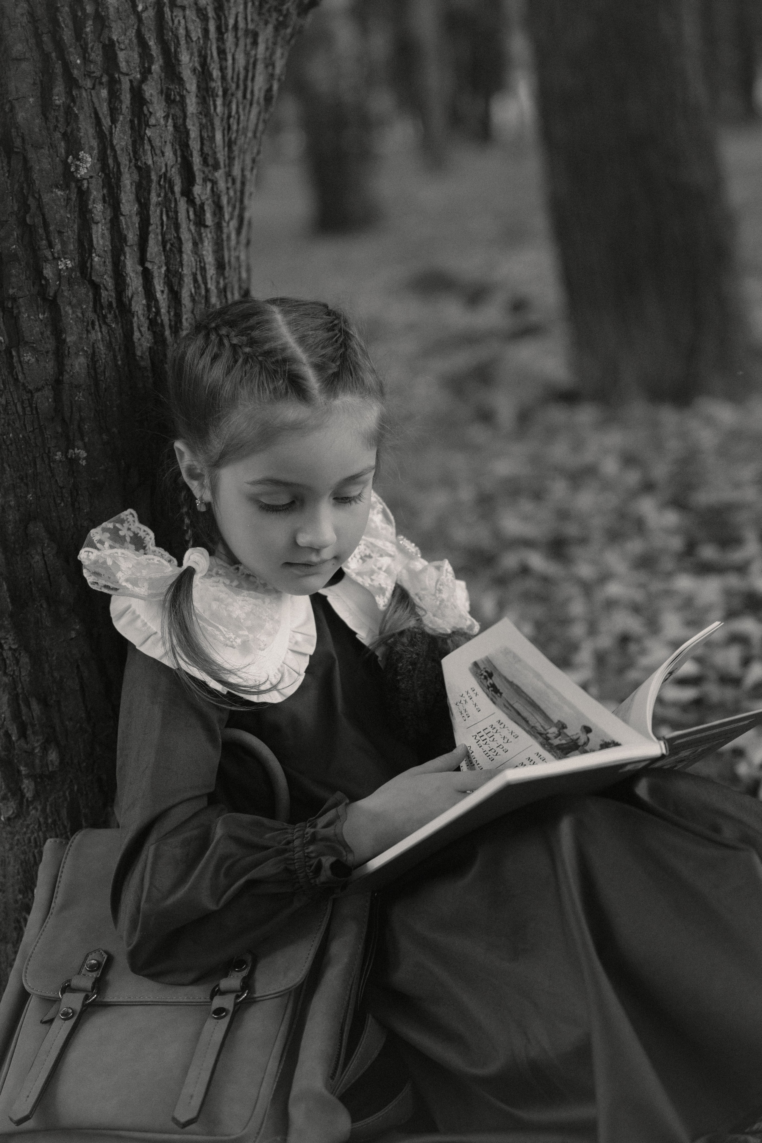 черно-белая фотография девочки, которая сидит в парке у дерева и читает букварь