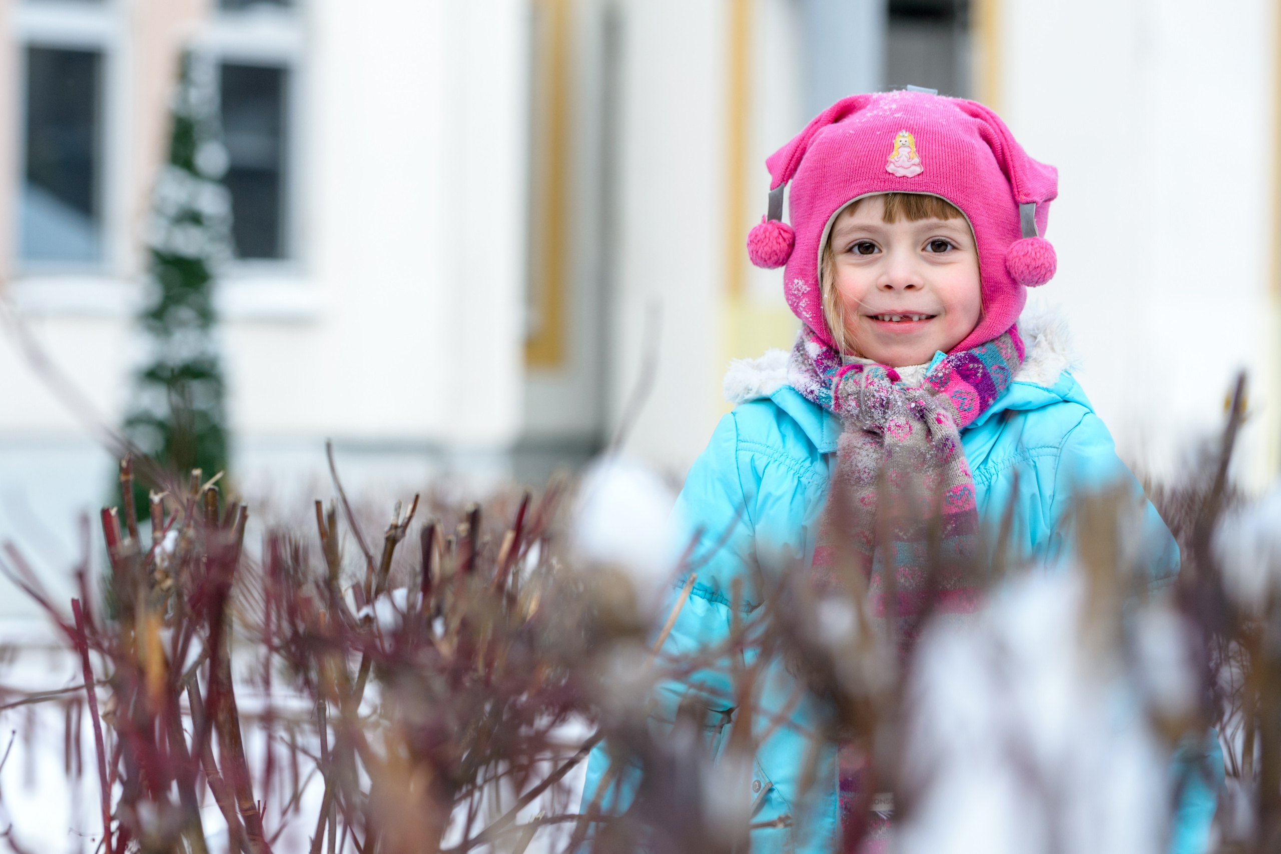Девочка за ветками деревьев в розовой шапке и голубой куртке улыбается.