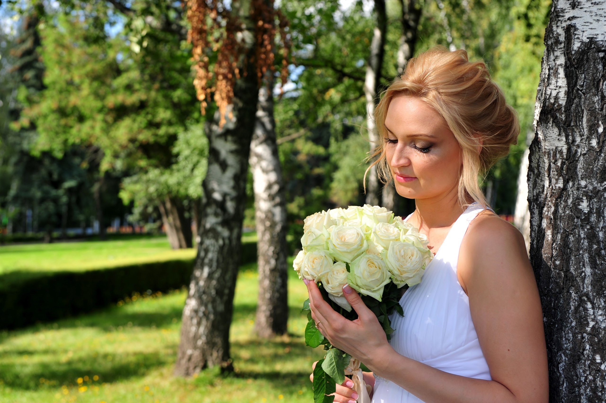 Свадьба на Воробьёвых Горах рядом с МГУ, невеста стоит, облокотившись спиной на берёзку в берёзовой роще рядом с МГУ, в руках невеста держит свадебный букет из кремовых роз и смотрит на букет