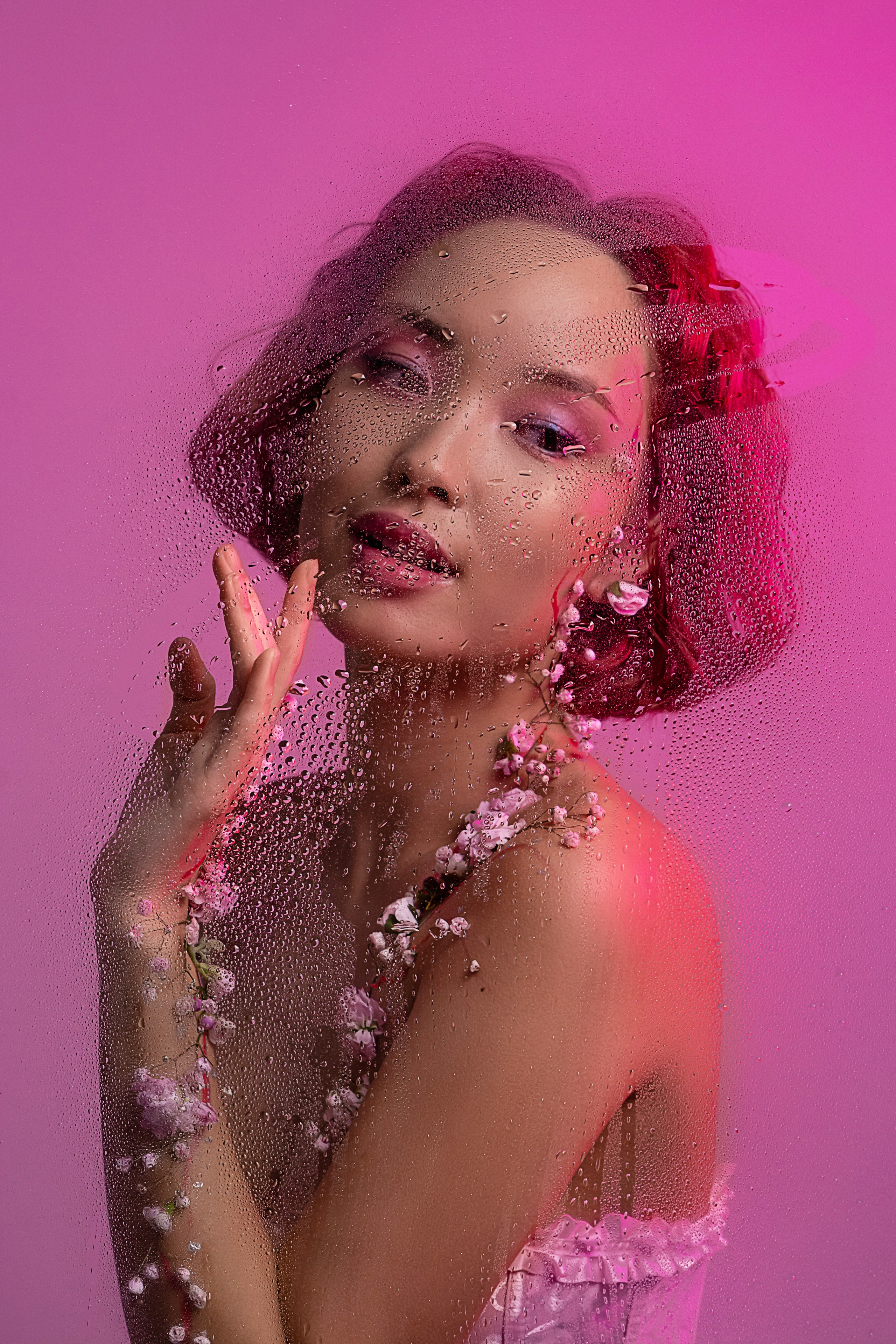 Мечтательный взгляд прекрасной азиатской девушки, чье тело украшено цветами, устремлен сквозь запотевшее стекло с каплями, символизируя ожидание весны.