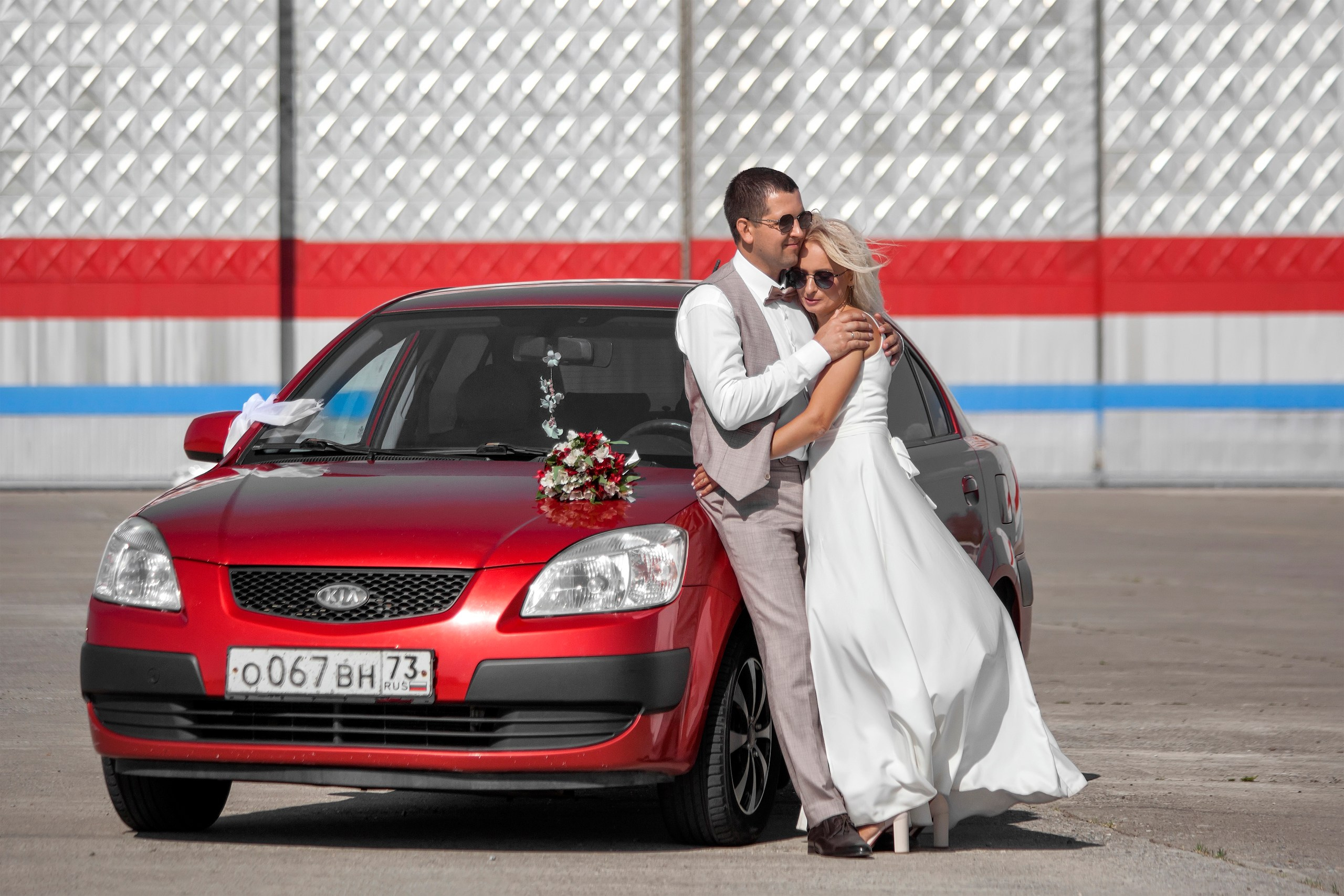 Жених с невестой обнимаются у красной машины в городской локации - свадебная фотосессия