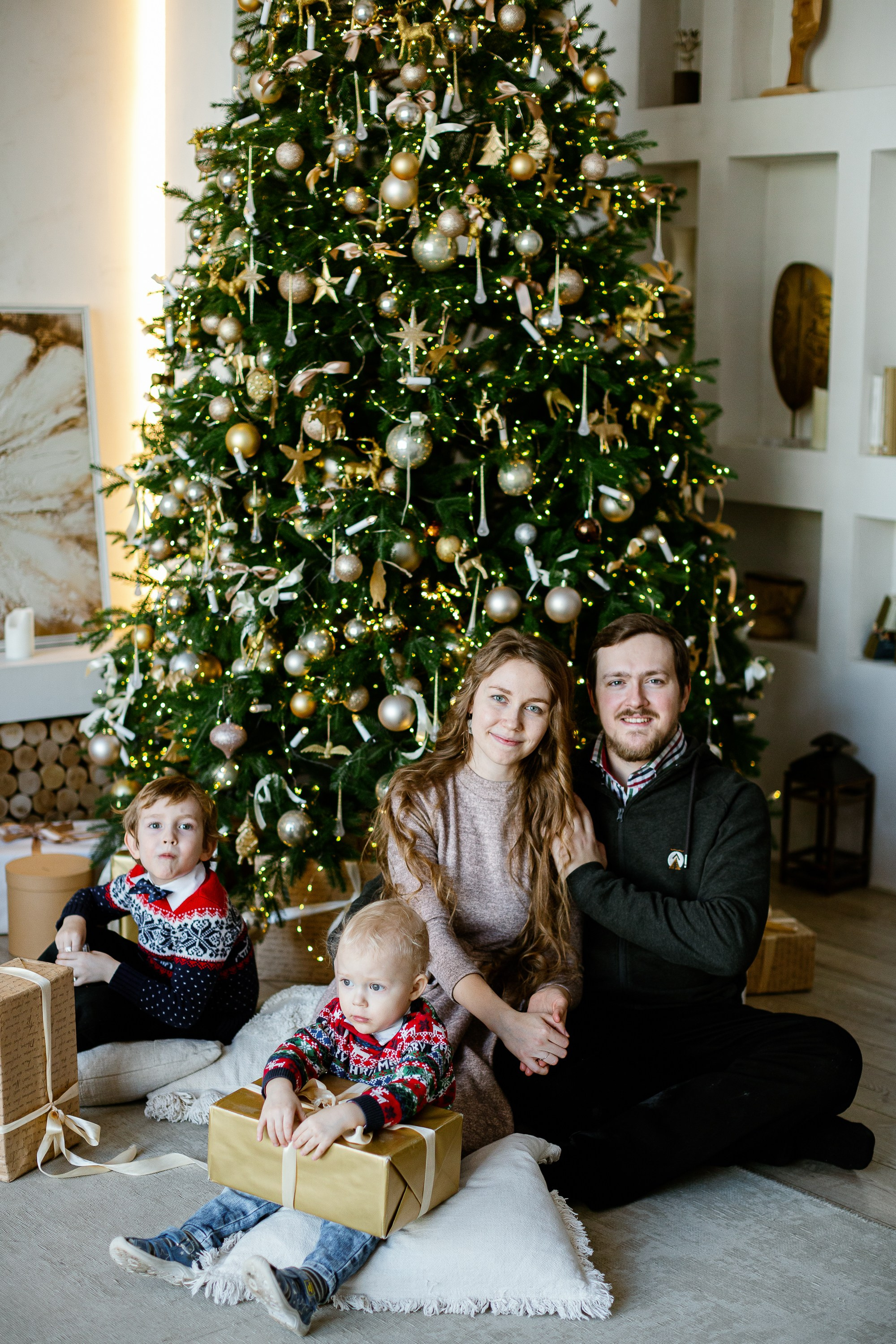 Семейная сидит на полу на фоне стоит елка и светится огоньками. Все улыбаются и смеются.
