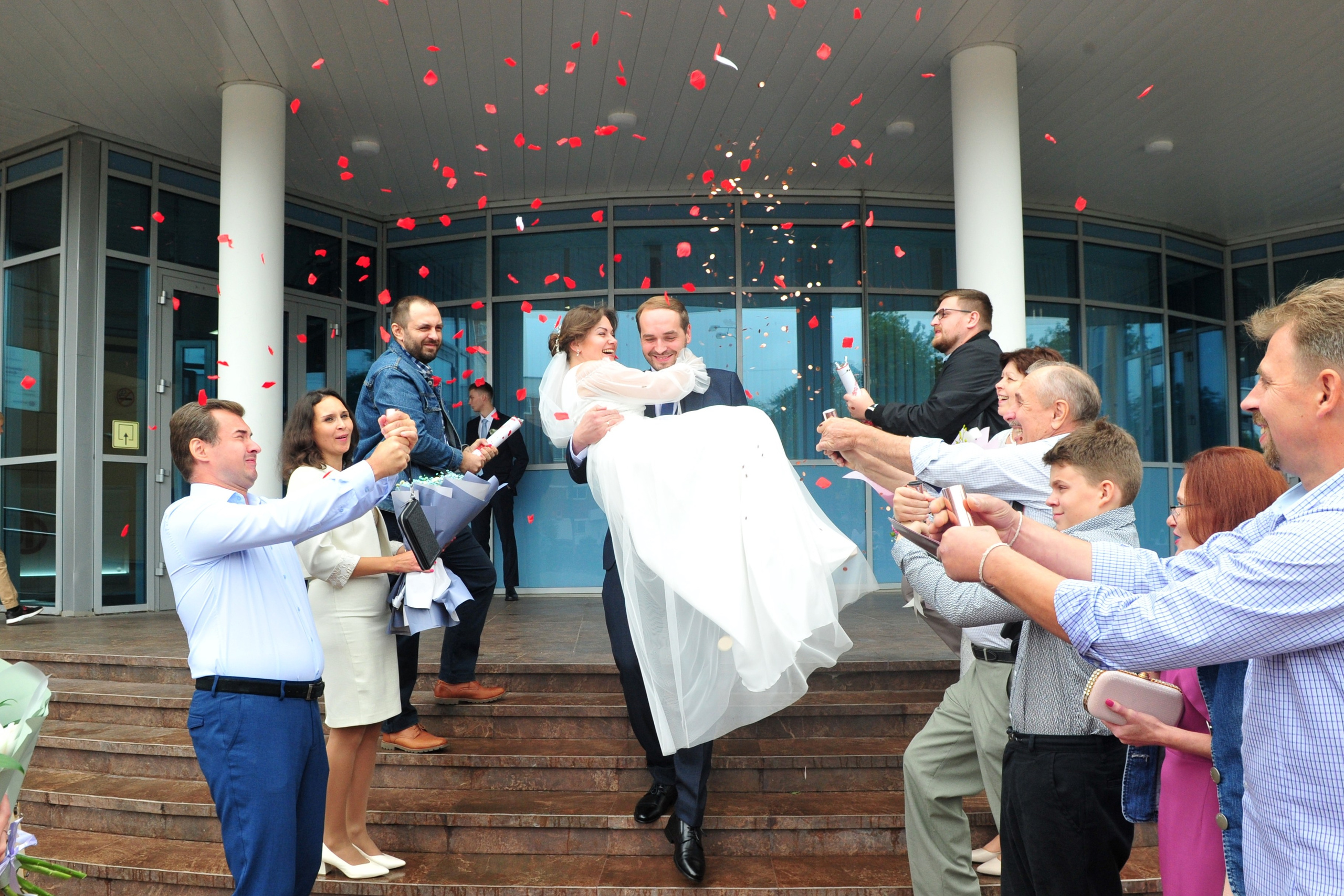 Свадьба в Царицыно пасмурным летним днем, жених выносит невесту на руках из Шипиловского ЗАГСа на фоне летящих лепестков и гостей