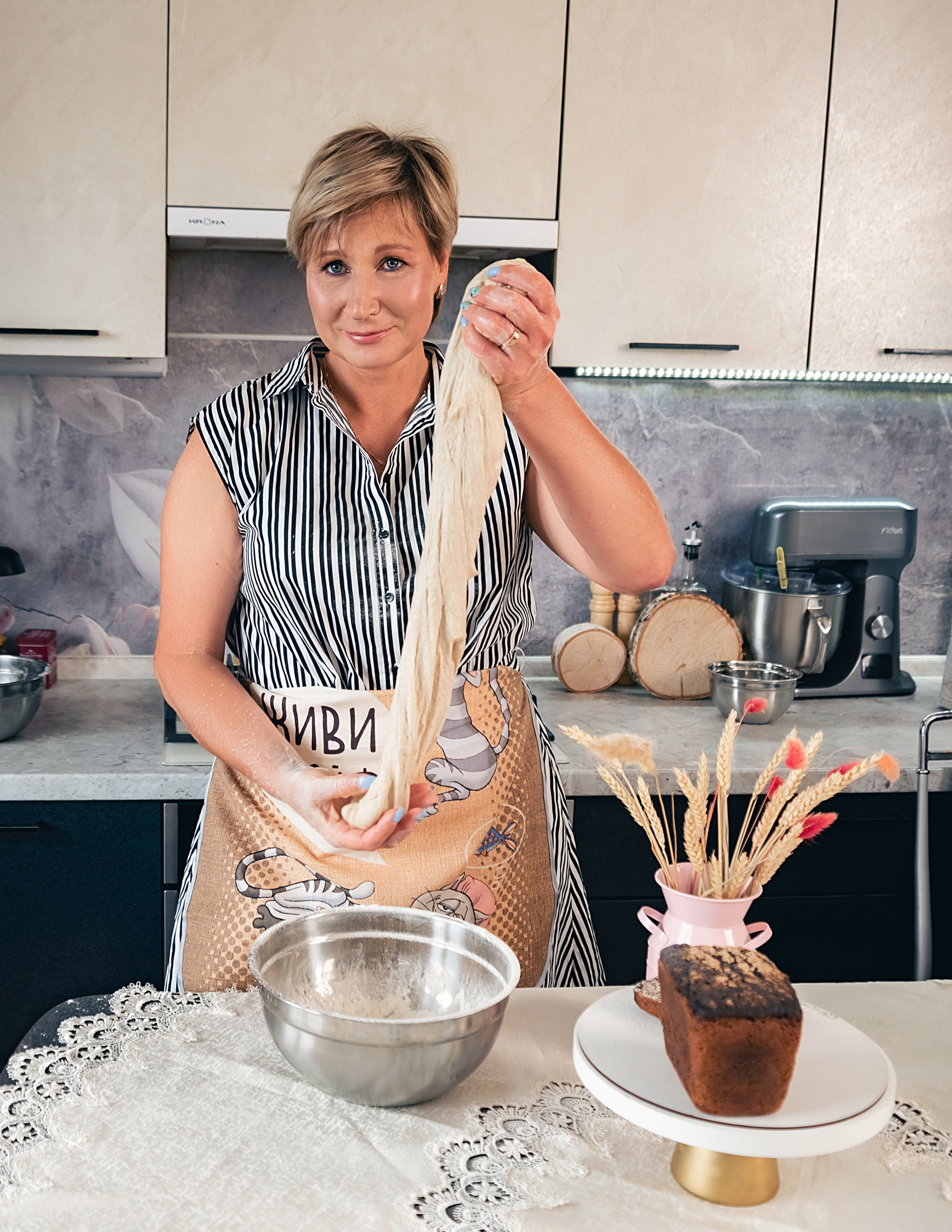 На фото женщина на кухне месит тесто.