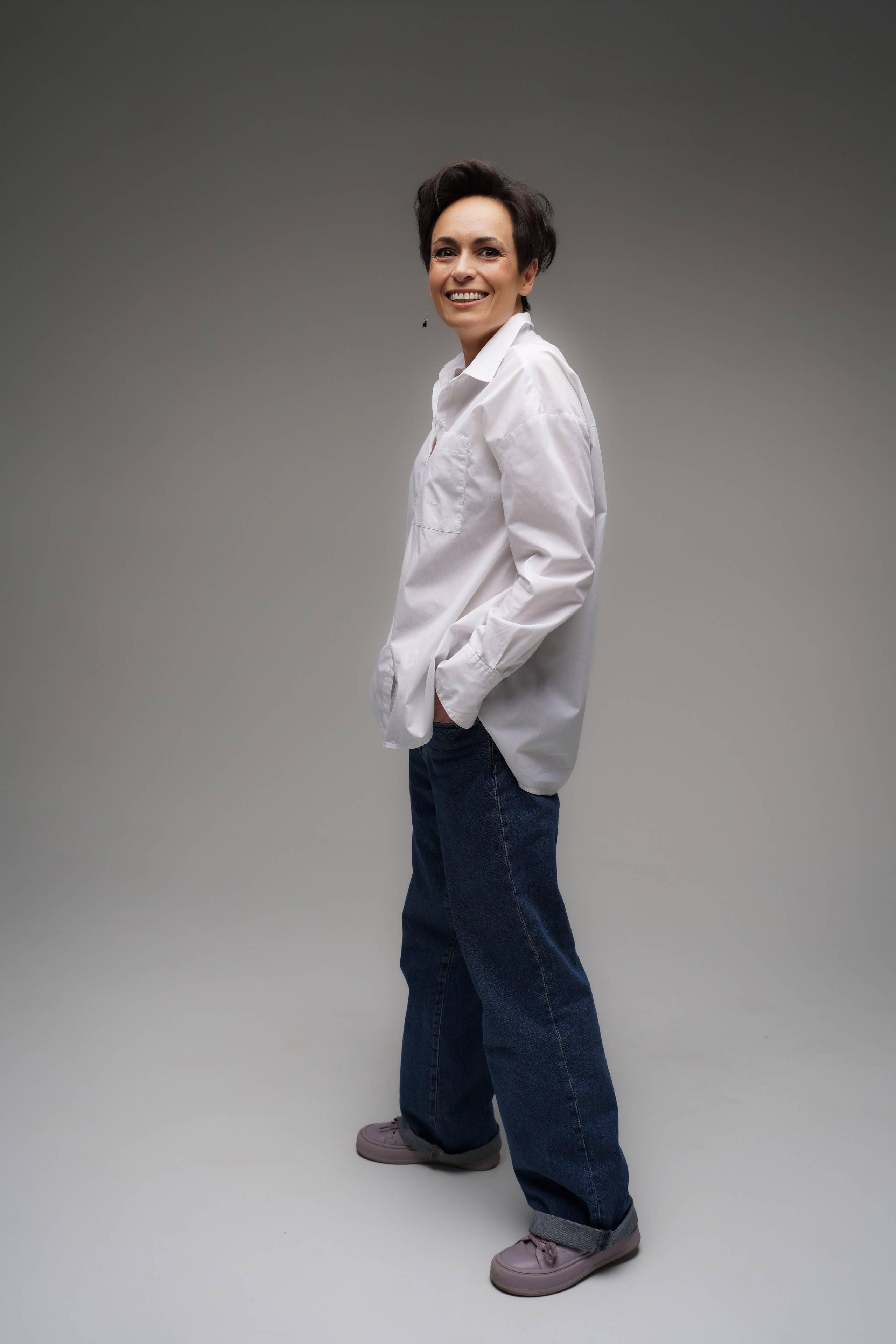 Девушка улыбается фотографу в фотостудии. Ее руки спрятаны в карманы 